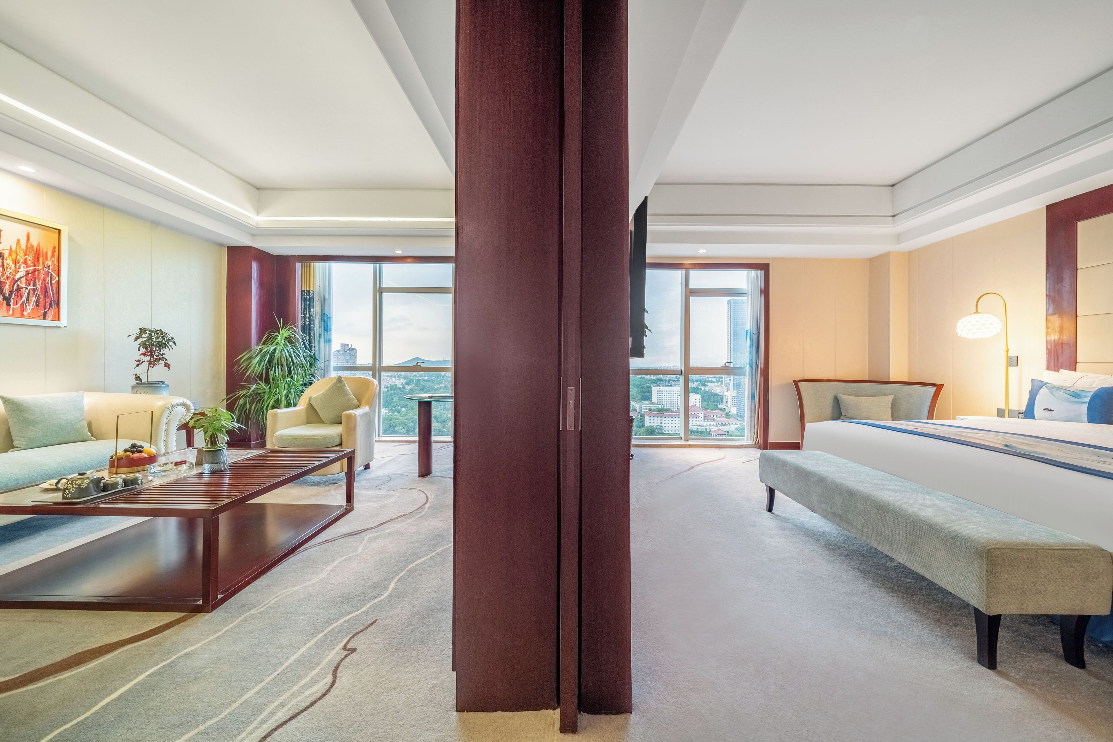 A modern hotel room with a sofa, a bench, a coffee table, and a large window offering a city view.