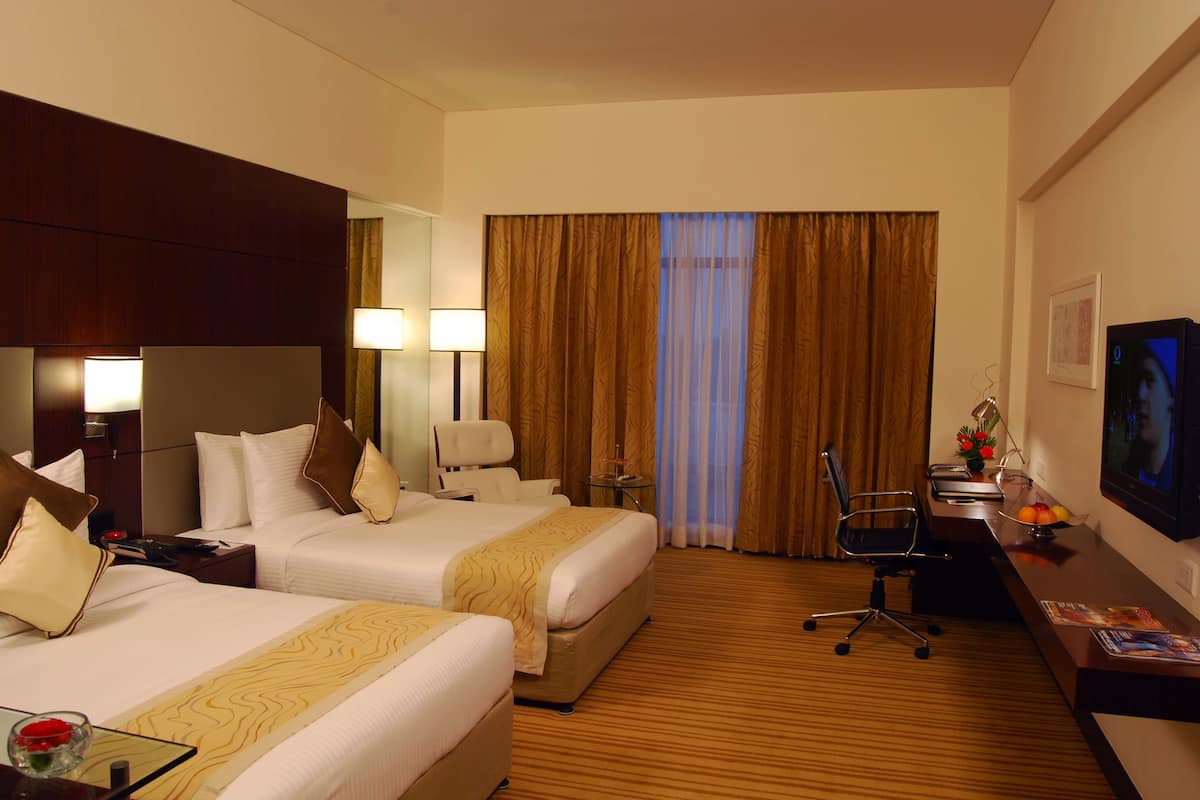 A hotel room with two beds, a desk, a chair, a TV, and a window with curtains.