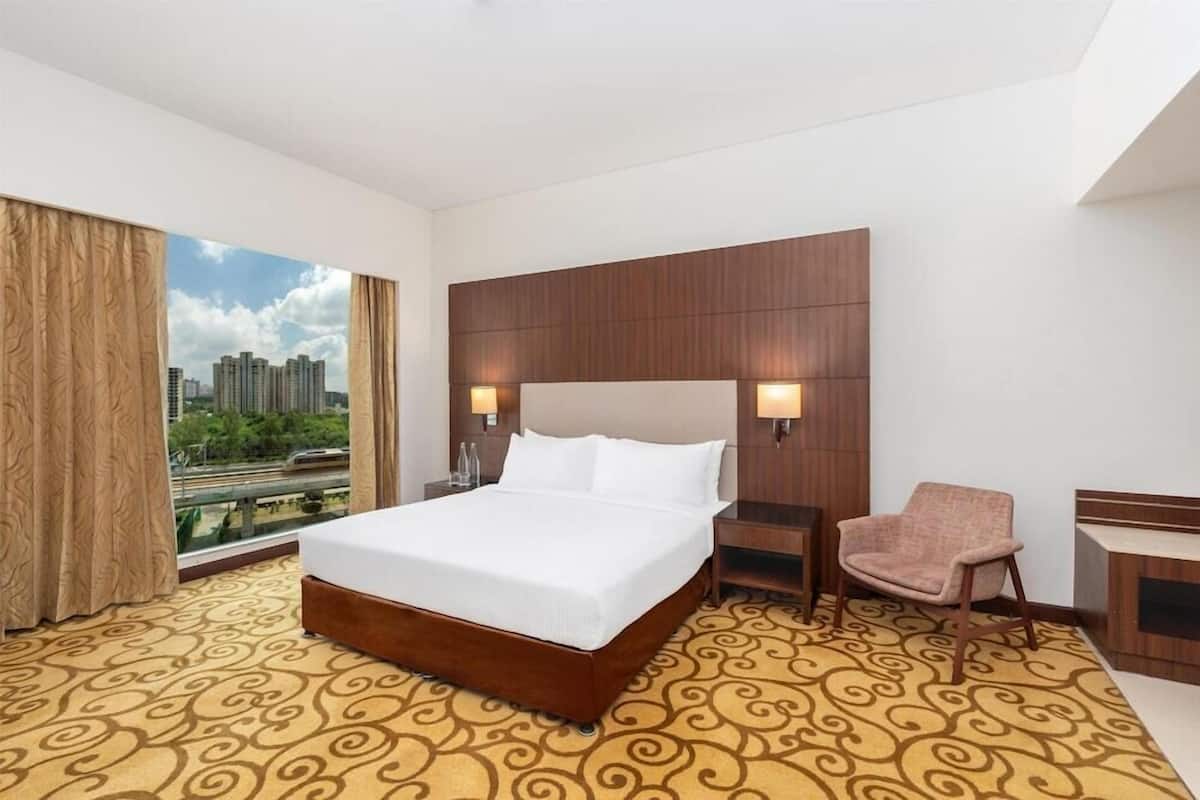 A hotel room with a large bed, a chair, a desk, and a view of buildings through a window.