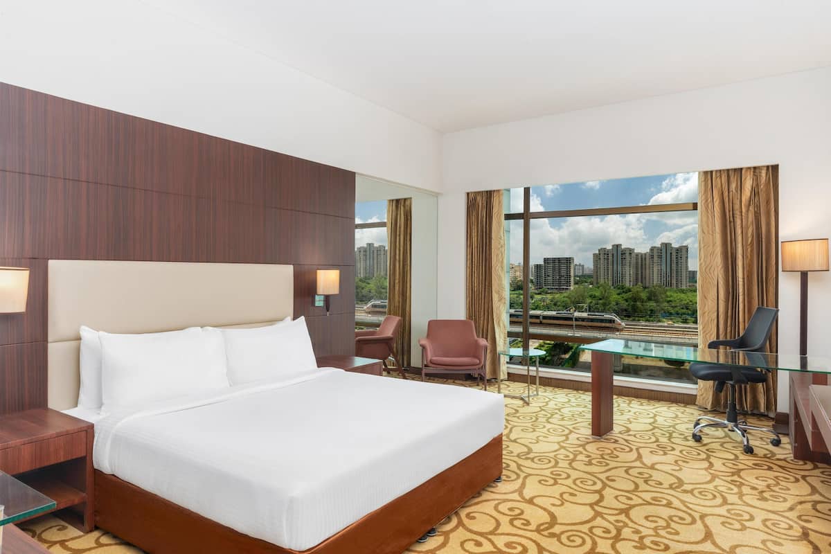 A hotel room with a large bed, a desk, a chair, and a view of a cityscape through large windows.
