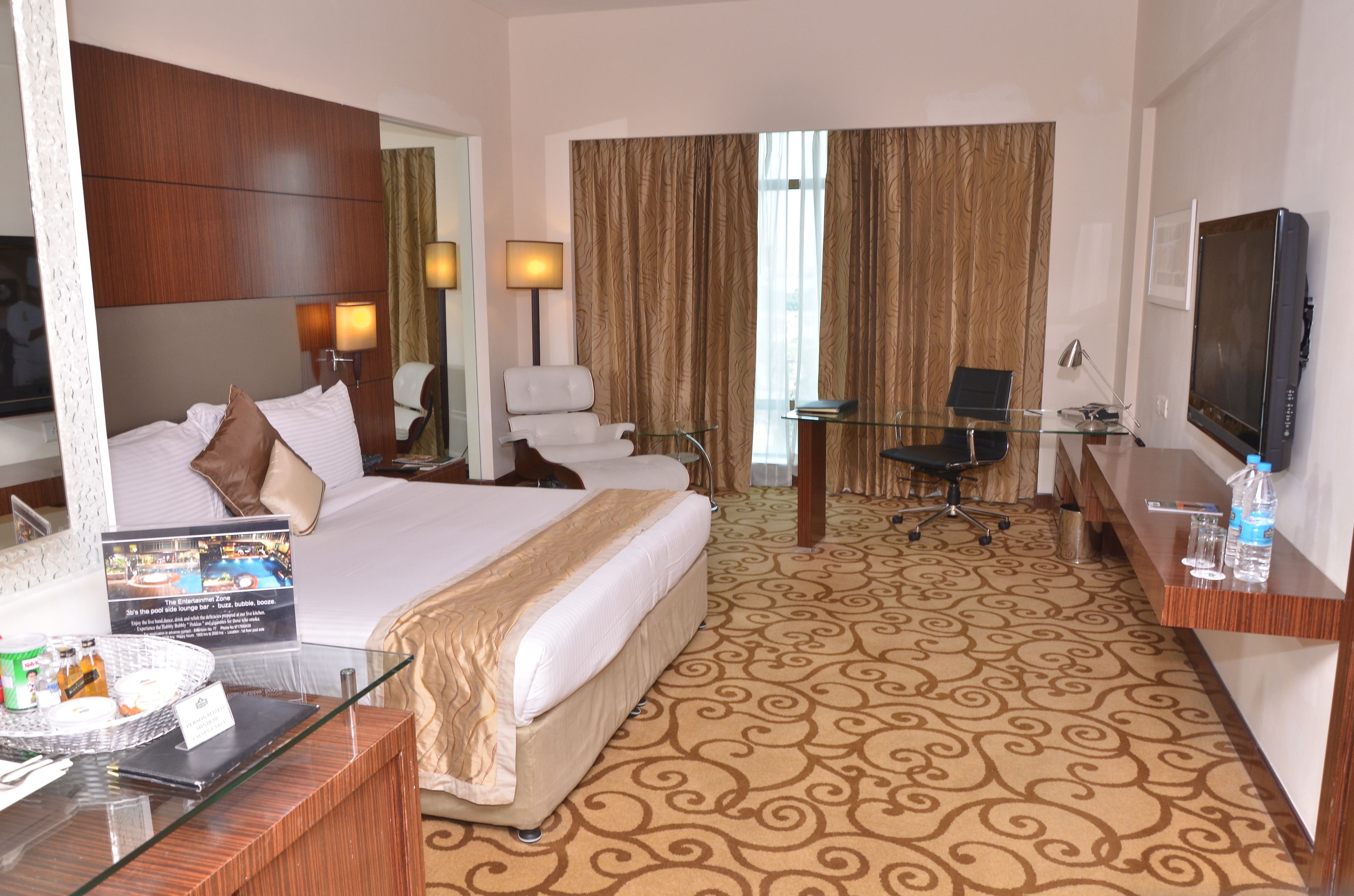 A hotel room with a large bed, a desk with a chair, a television, and a view of the city through curtains.