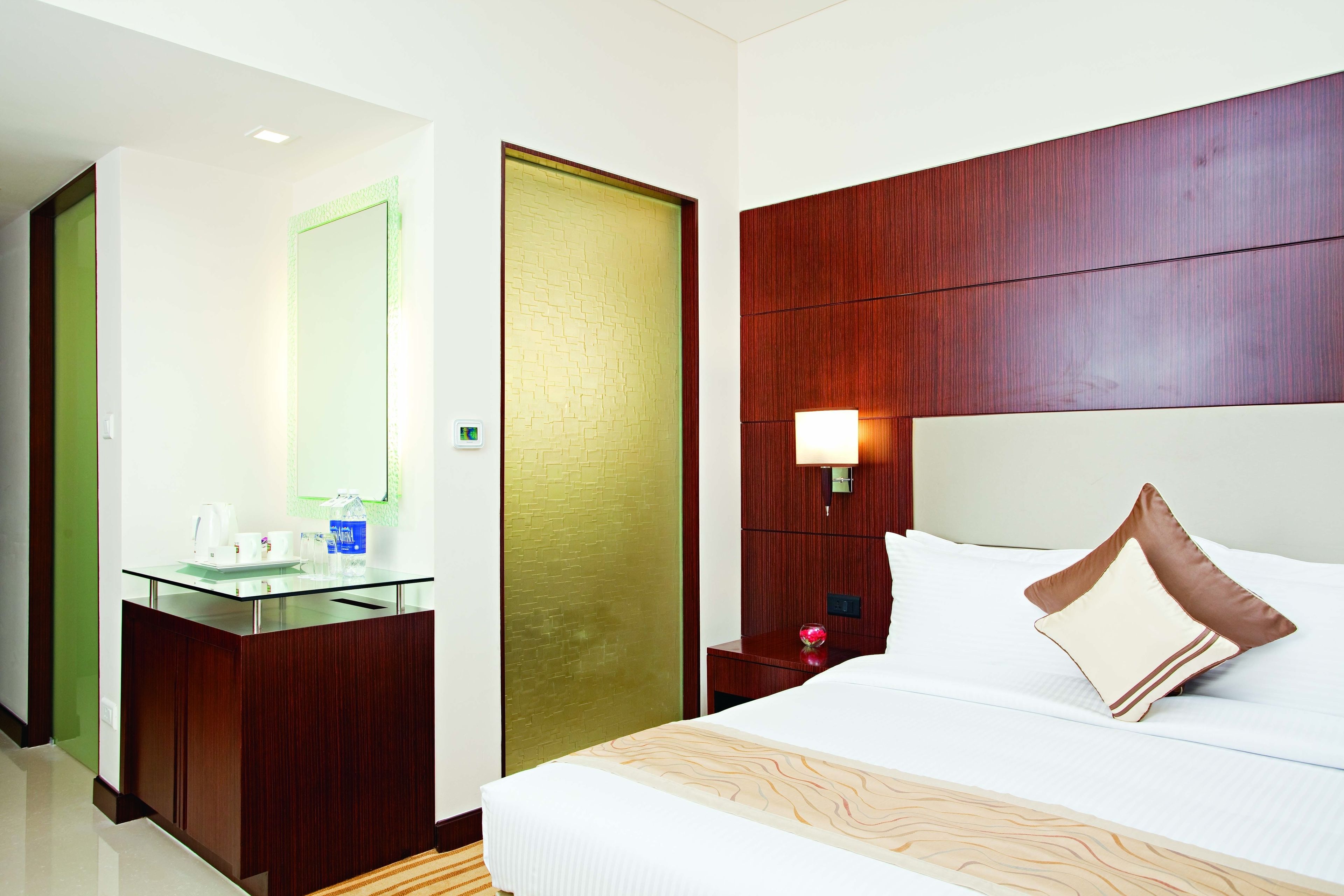 Una habitación de hotel con cabecera de madera, mesita de noche de vidrio, cama con ropa de cama blanca y mesita con lámpara.