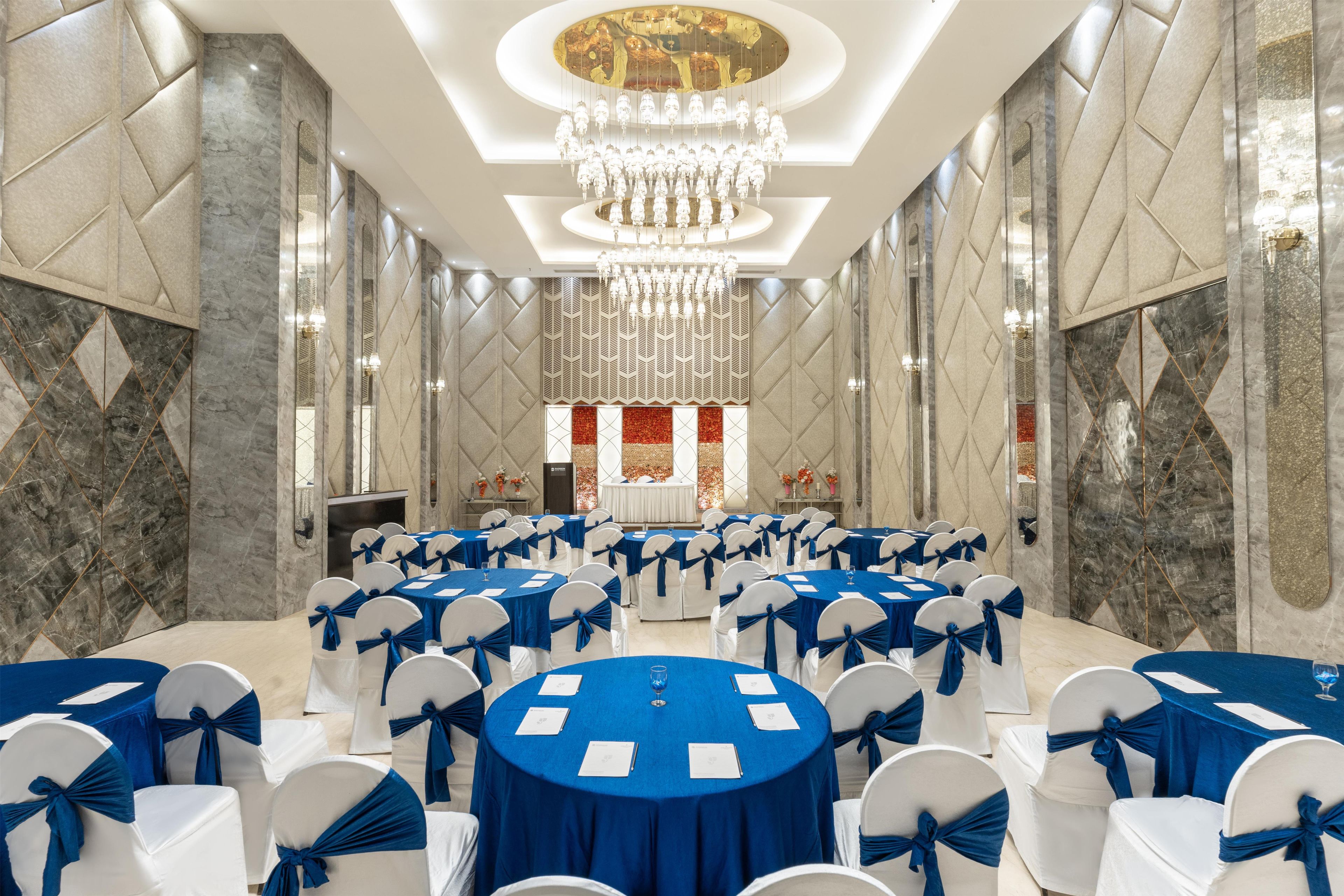 A banquet hall with round tables, white chairs with blue sashes, and a large chandelier.