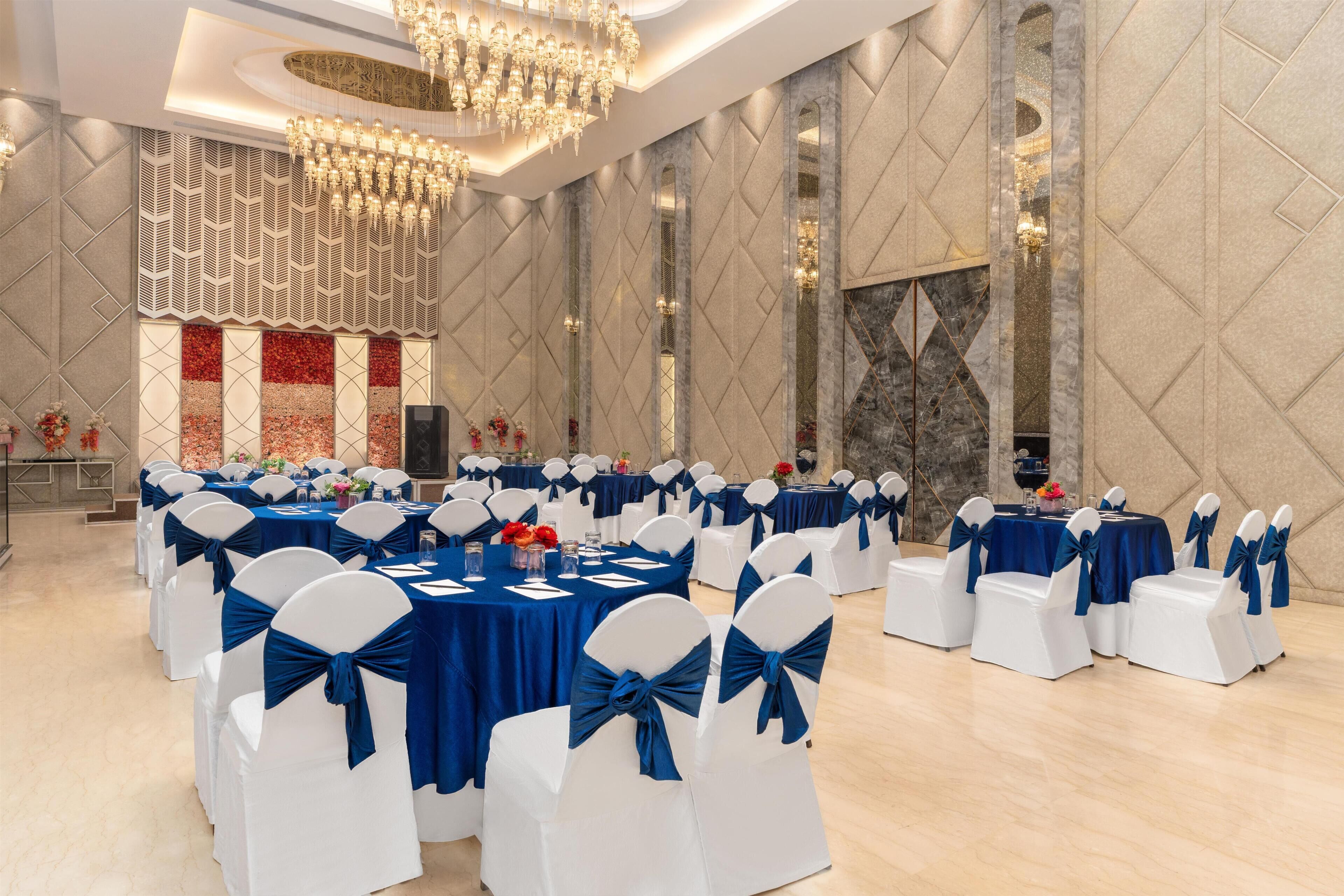 A banquet hall with round tables, white chairs with blue sashes, and a decorative wall with a geometric pattern.