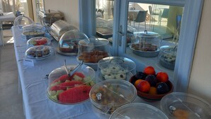 A buffet table with various food items, including fruits and desserts, in a room with large windows and a view of the sea. - Kedros Villas (Naxos)