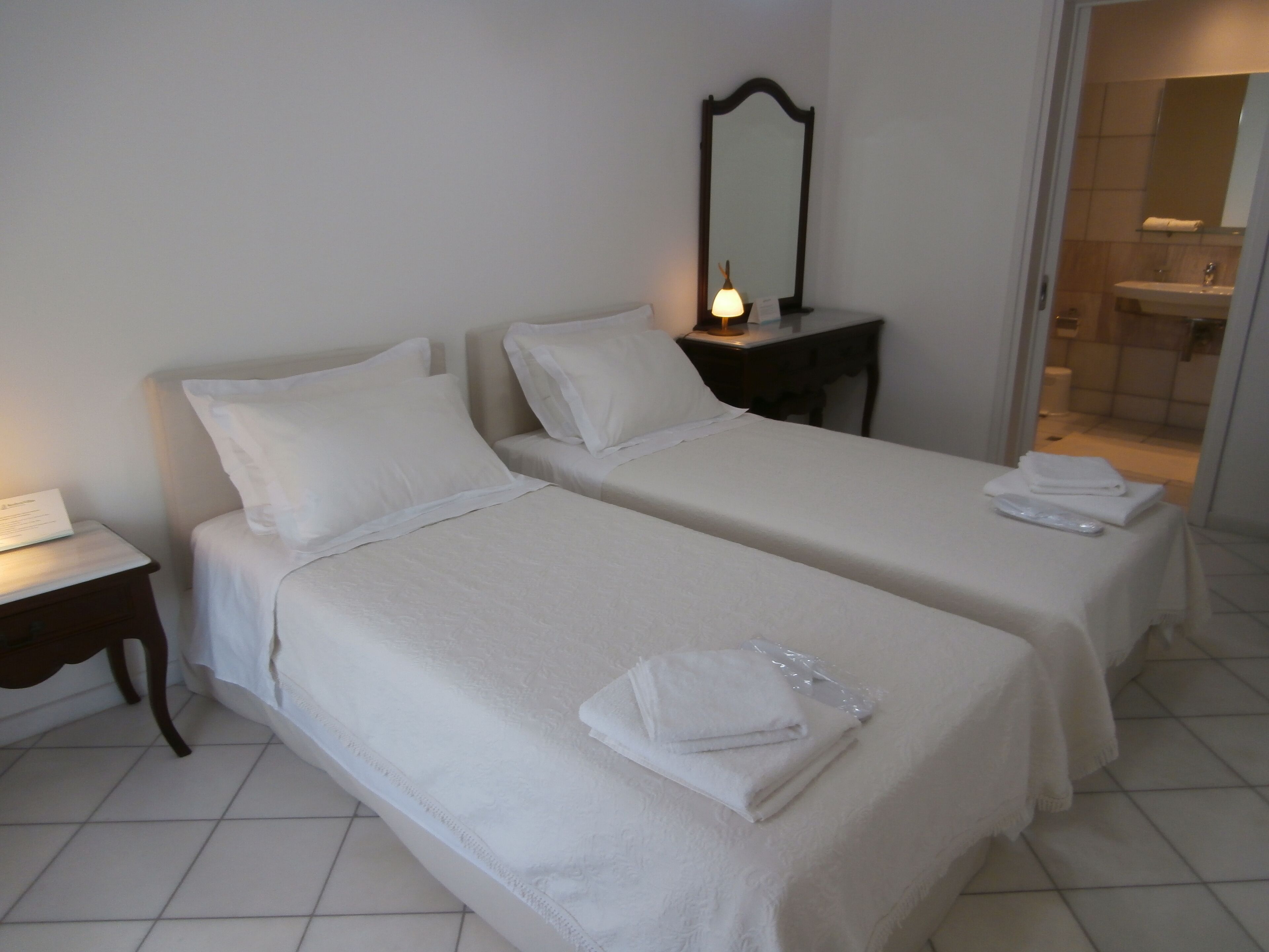 A hotel room with two beds, a bedside table with a lamp, a mirror, and a bathroom visible through an open door.