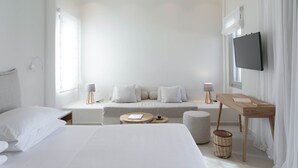 A modern hotel room with a large bed, a sofa, a small round table, a TV, and a window with curtains. - Kedros Villas (Naxos)
