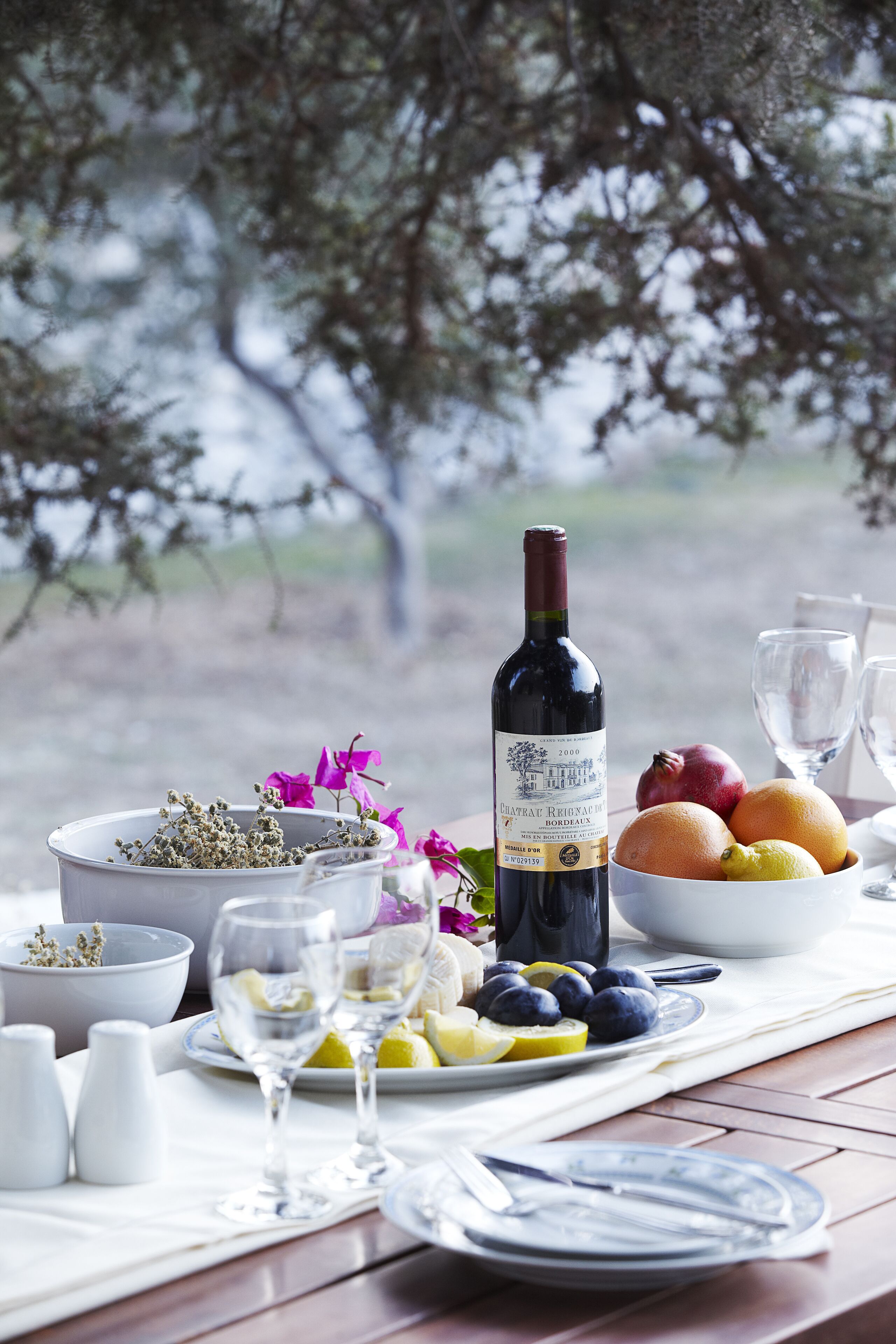 A table set with a bottle of wine, bowls of fruit, and glasses, with a view of trees in the background.