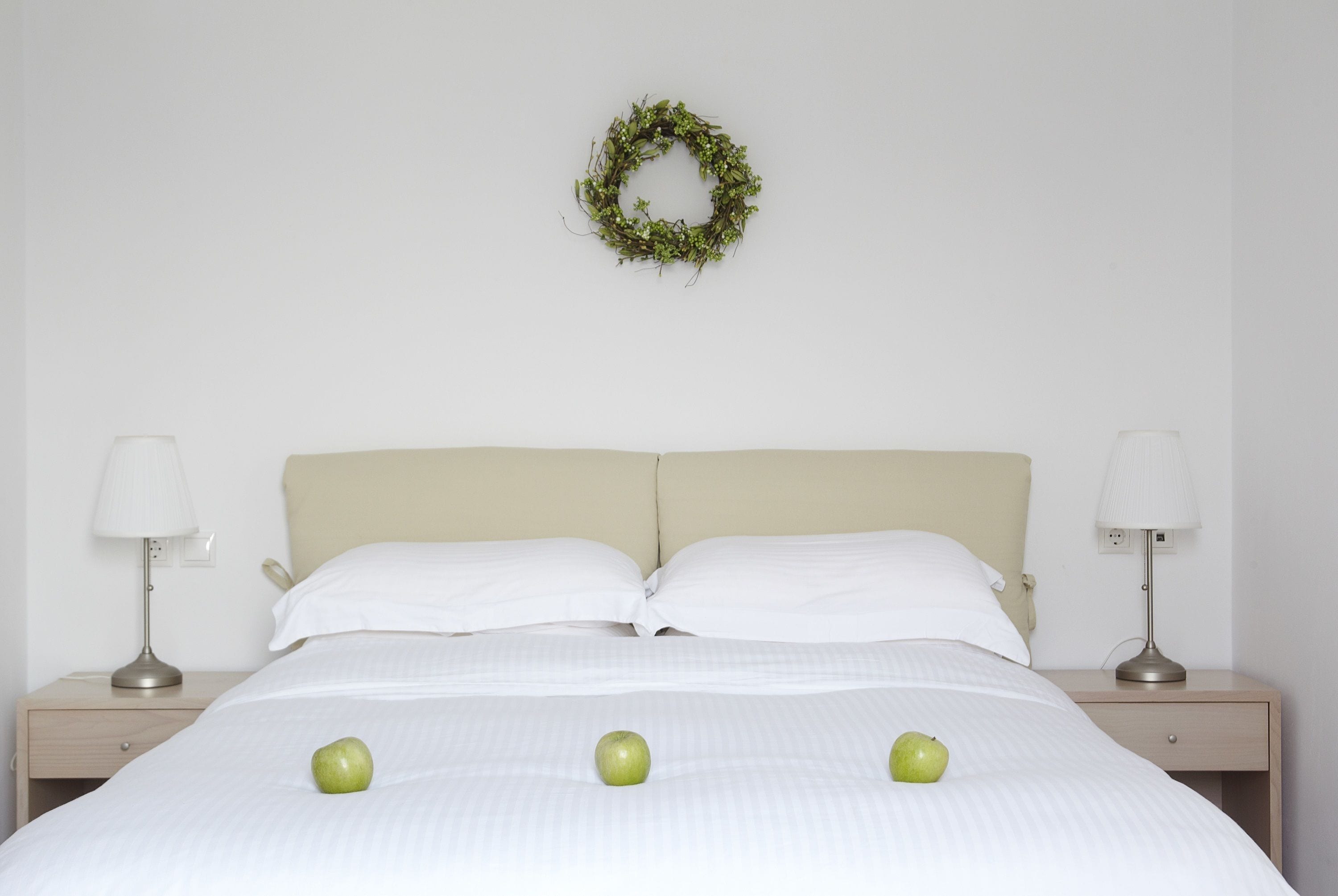 A neatly made bed with white linens and three green apples on it, flanked by two bedside tables with lamps, and a simple green wreath on the wall.