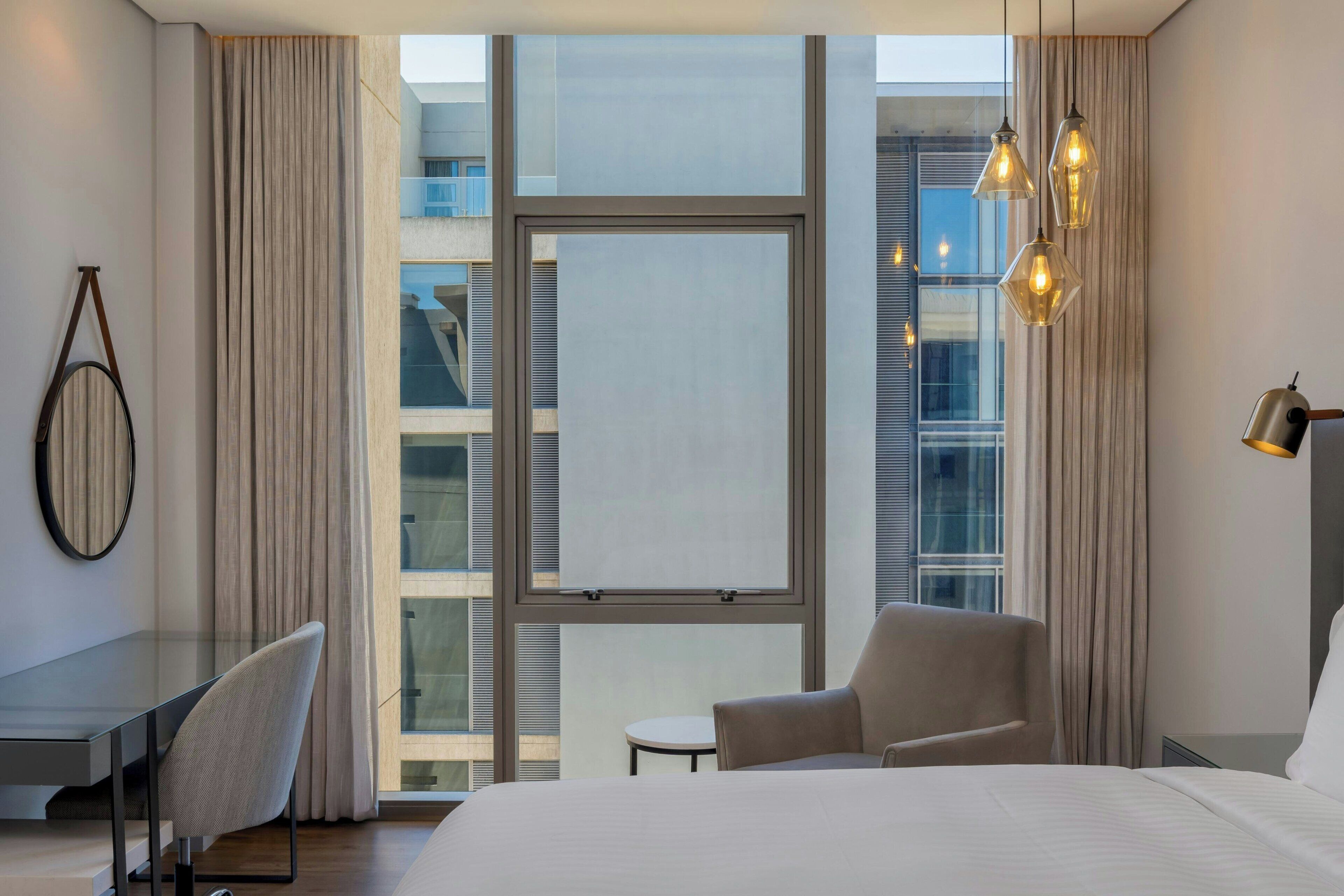 Habitación de hotel moderna con una cama grande, un escritorio y una silla. Cuenta con una ventana con cortinas y vistas a la ciudad.