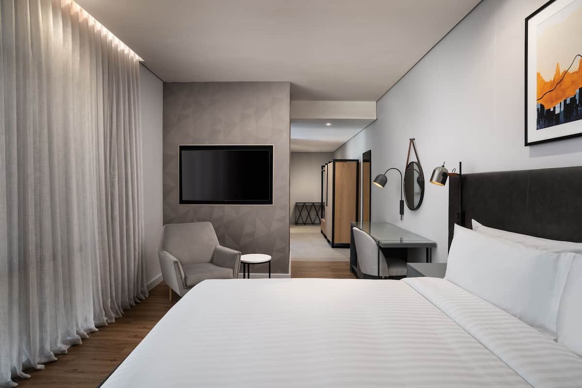 A modern hotel room with a large bed, a desk, a chair, a TV, and a painting on the wall.