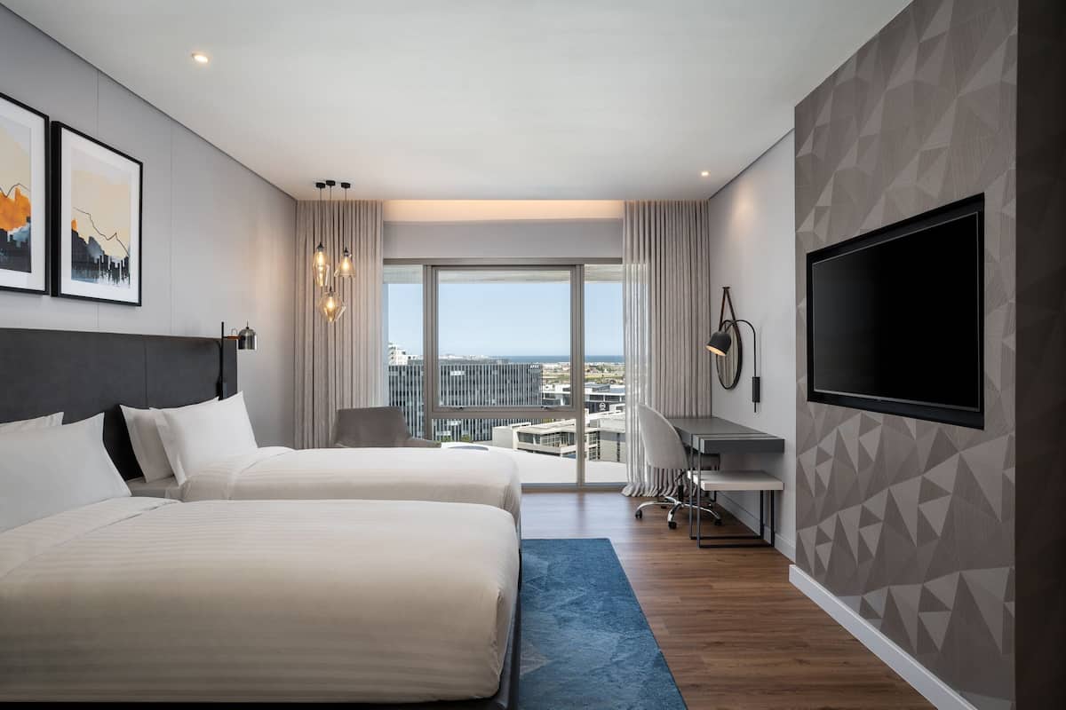 A modern hotel room with two beds, a desk, a TV, and a city view.