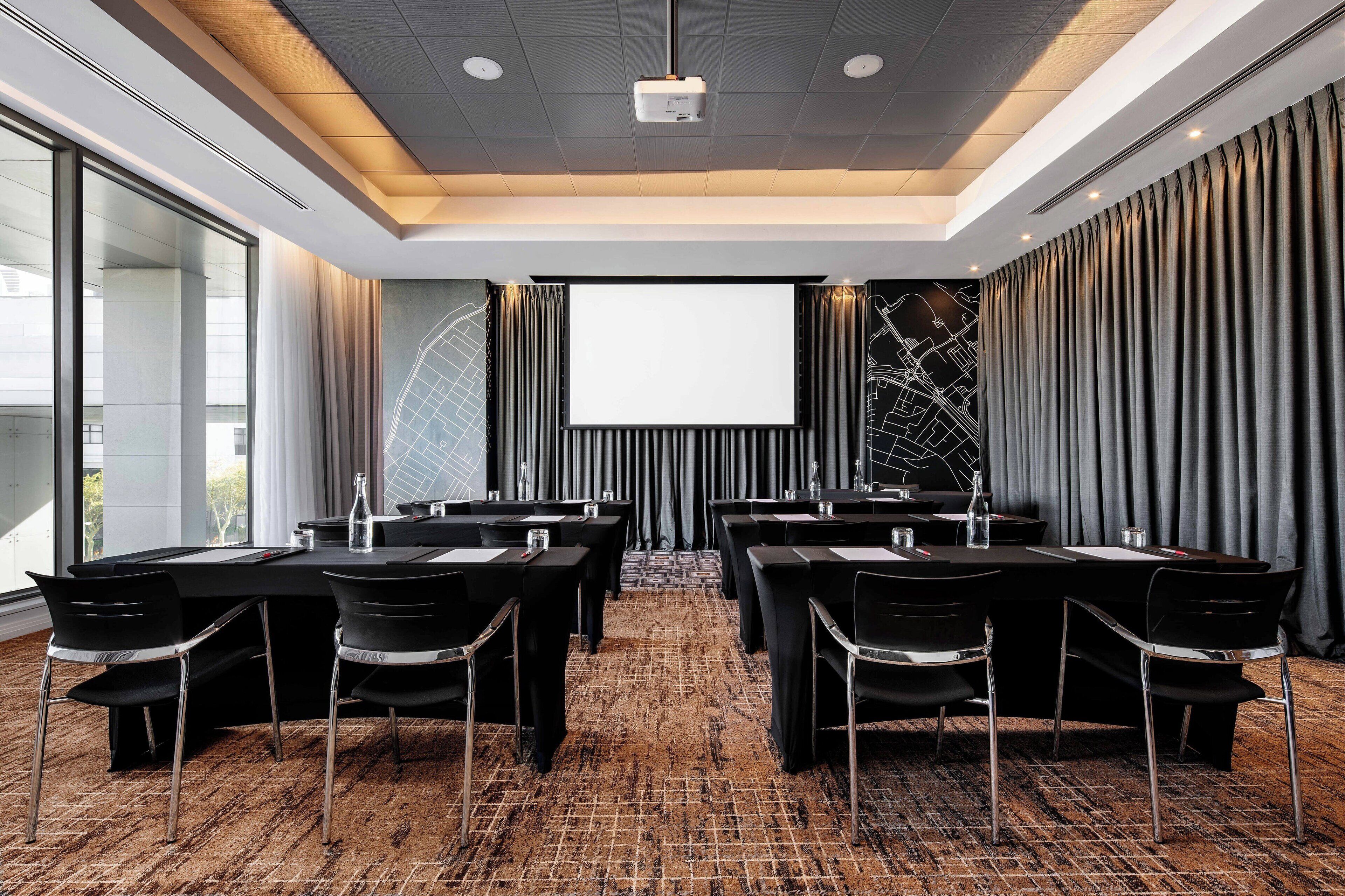 Un salón de conferencias con una pantalla grande, mesas y sillas negras, piso alfombrado y vistas a la ciudad.