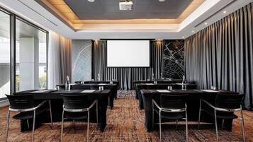Un salón de conferencias con una pantalla grande, mesas y sillas negras, piso alfombrado y vistas a la ciudad.
