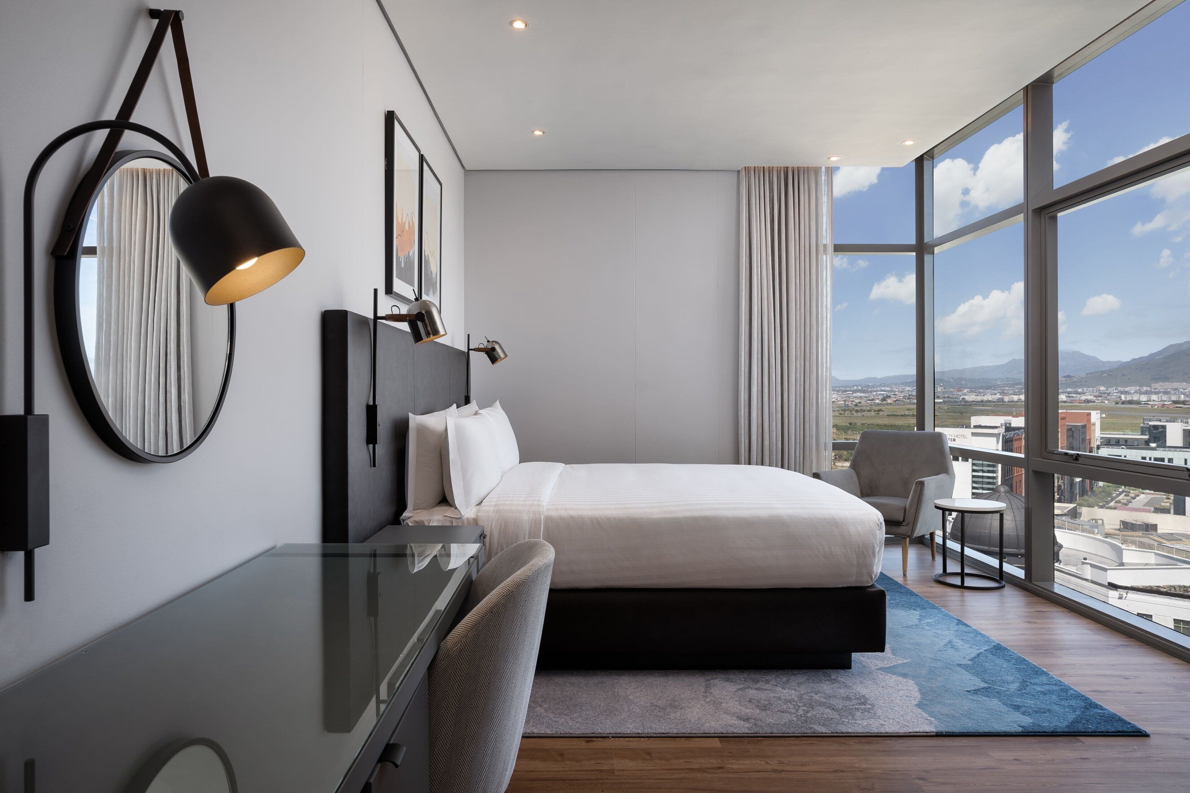 A modern hotel room with a large bed, a desk, a chair, a small table, and a view of the city through large windows.