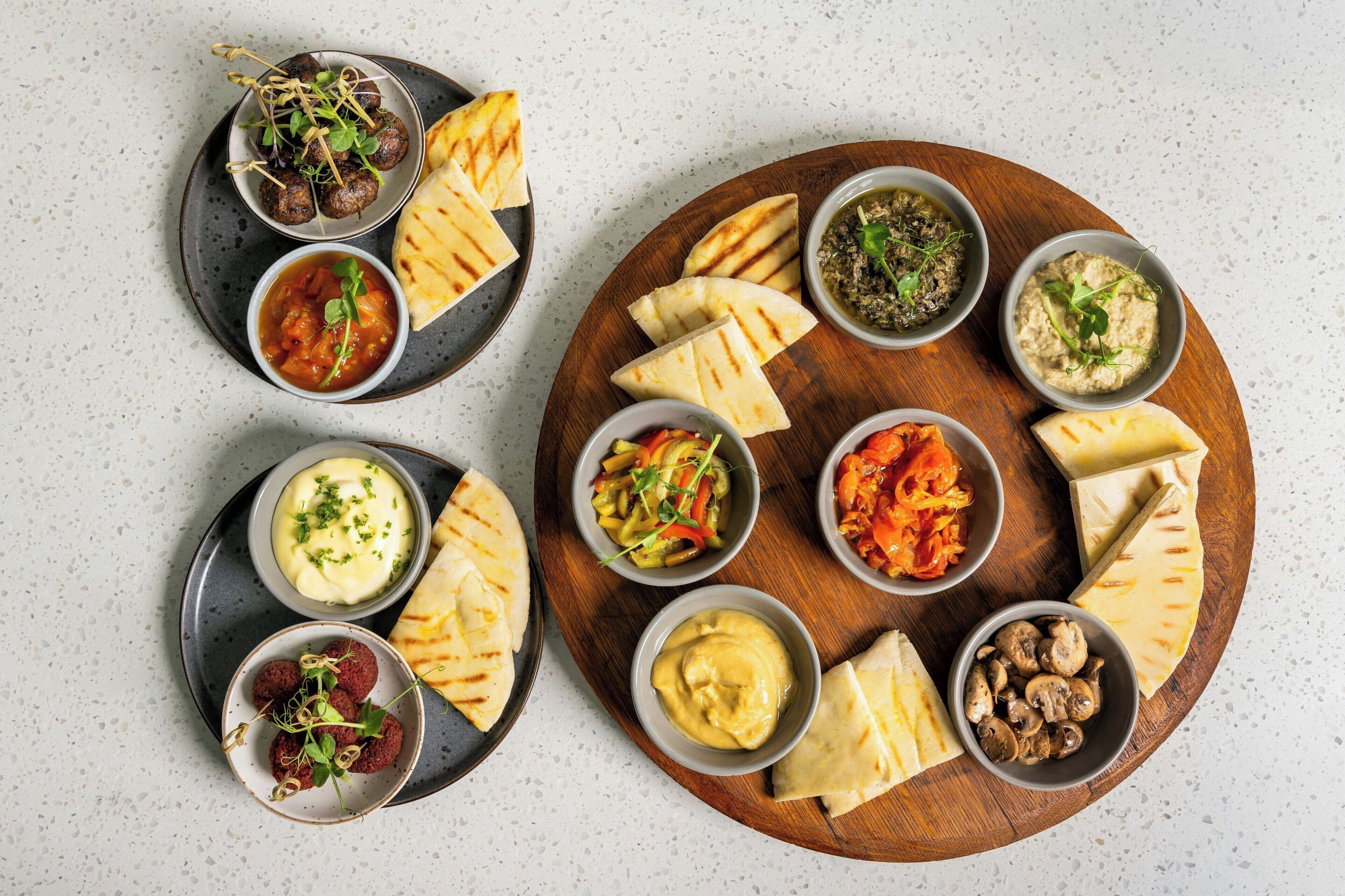 A wooden board with various food items and condiments, including grilled meatballs, flatbreads, and dips.