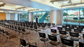 Un salón de conferencias con filas de sillas, ventanas amplias y vistas a un puente y palmeras.