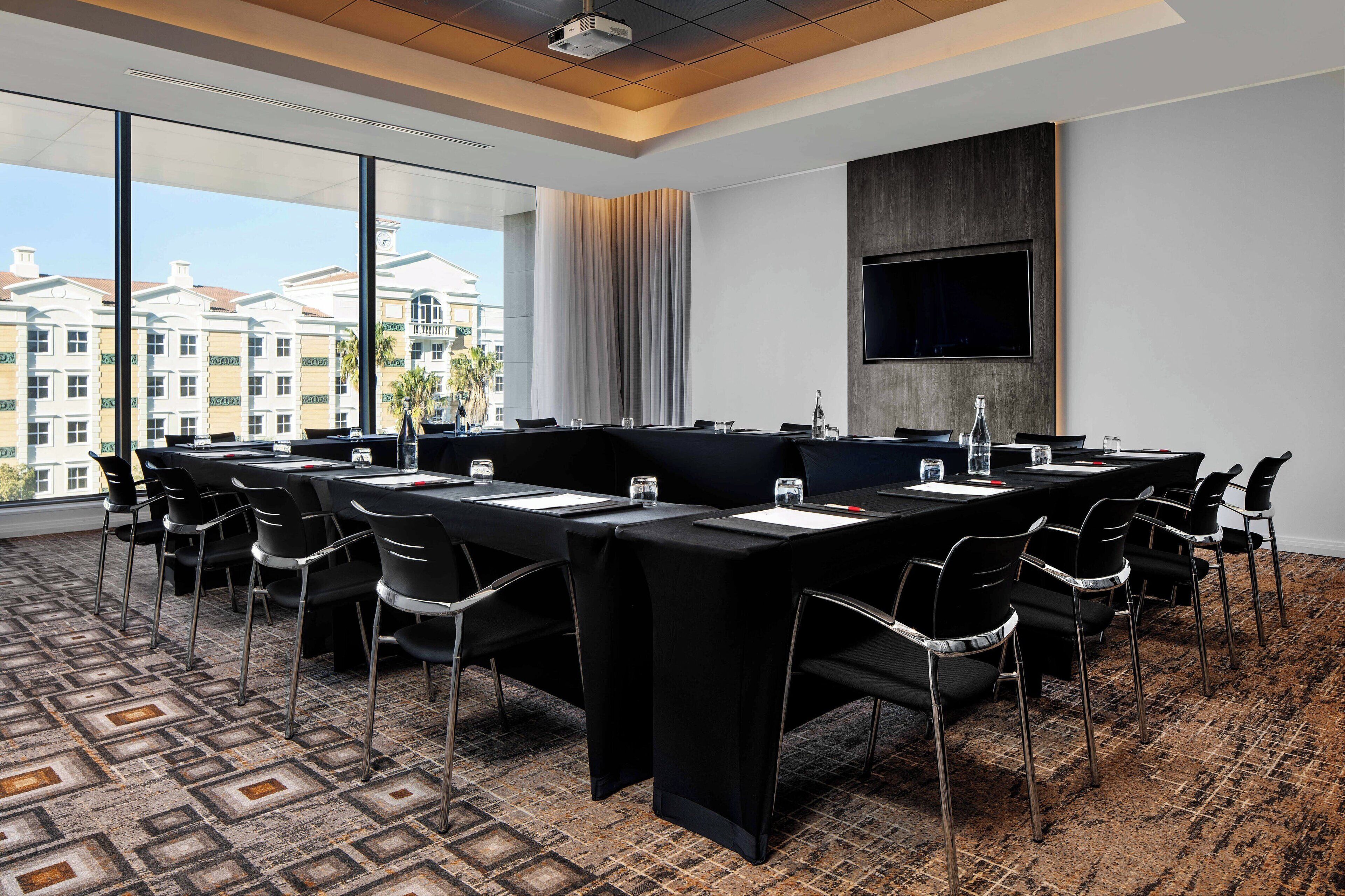 A conference room with a long table, chairs, a TV, and a large window with a city view.