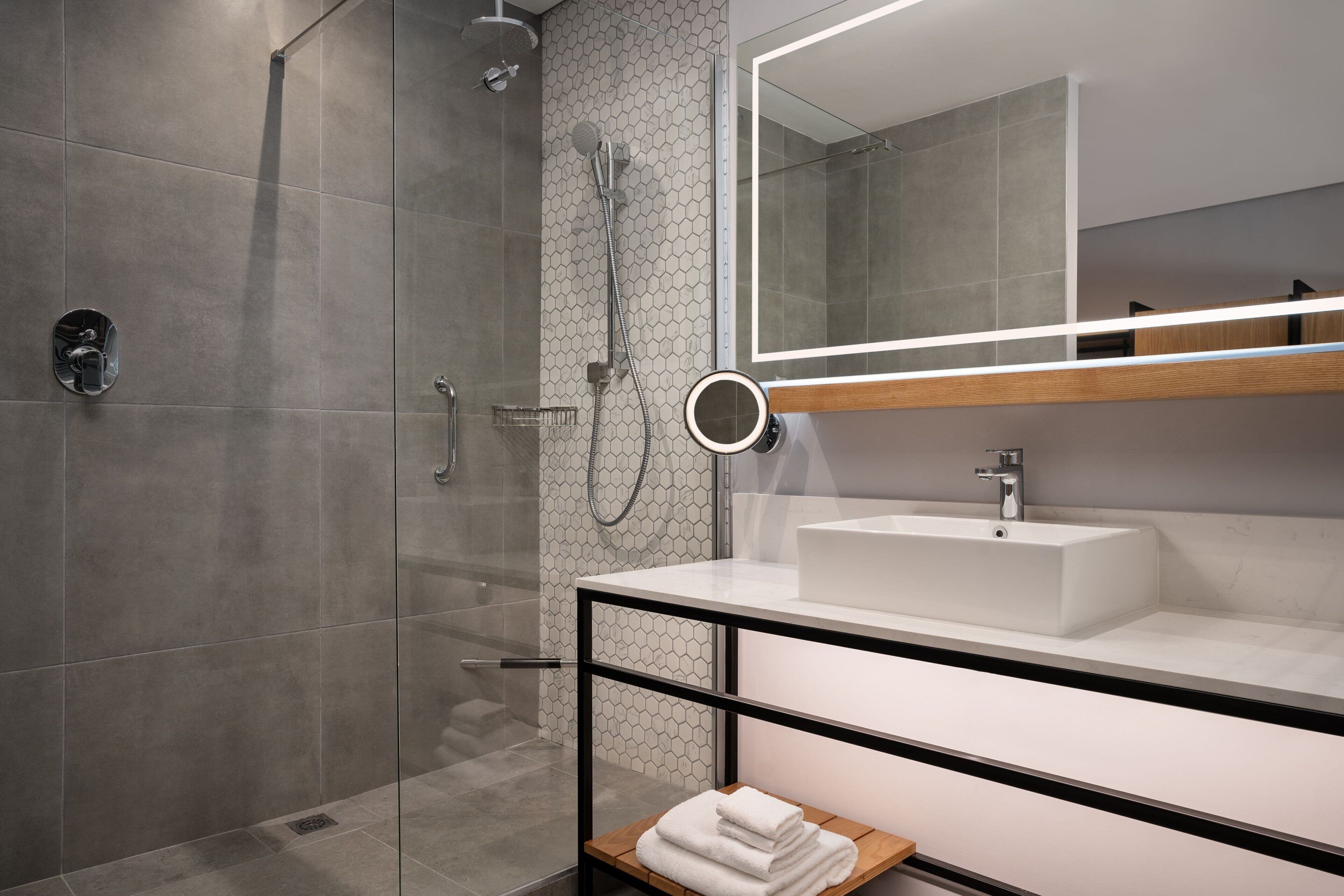 A modern bathroom with a glass-enclosed shower, a white sink, and a mirror with a wooden shelf above it.