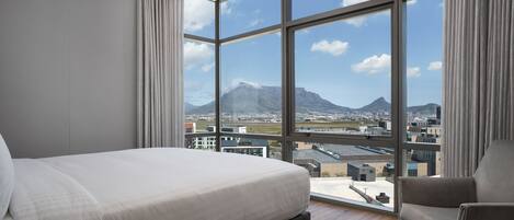 A modern bedroom with a large bed, a chair, and a view of a cityscape and mountains through large windows.