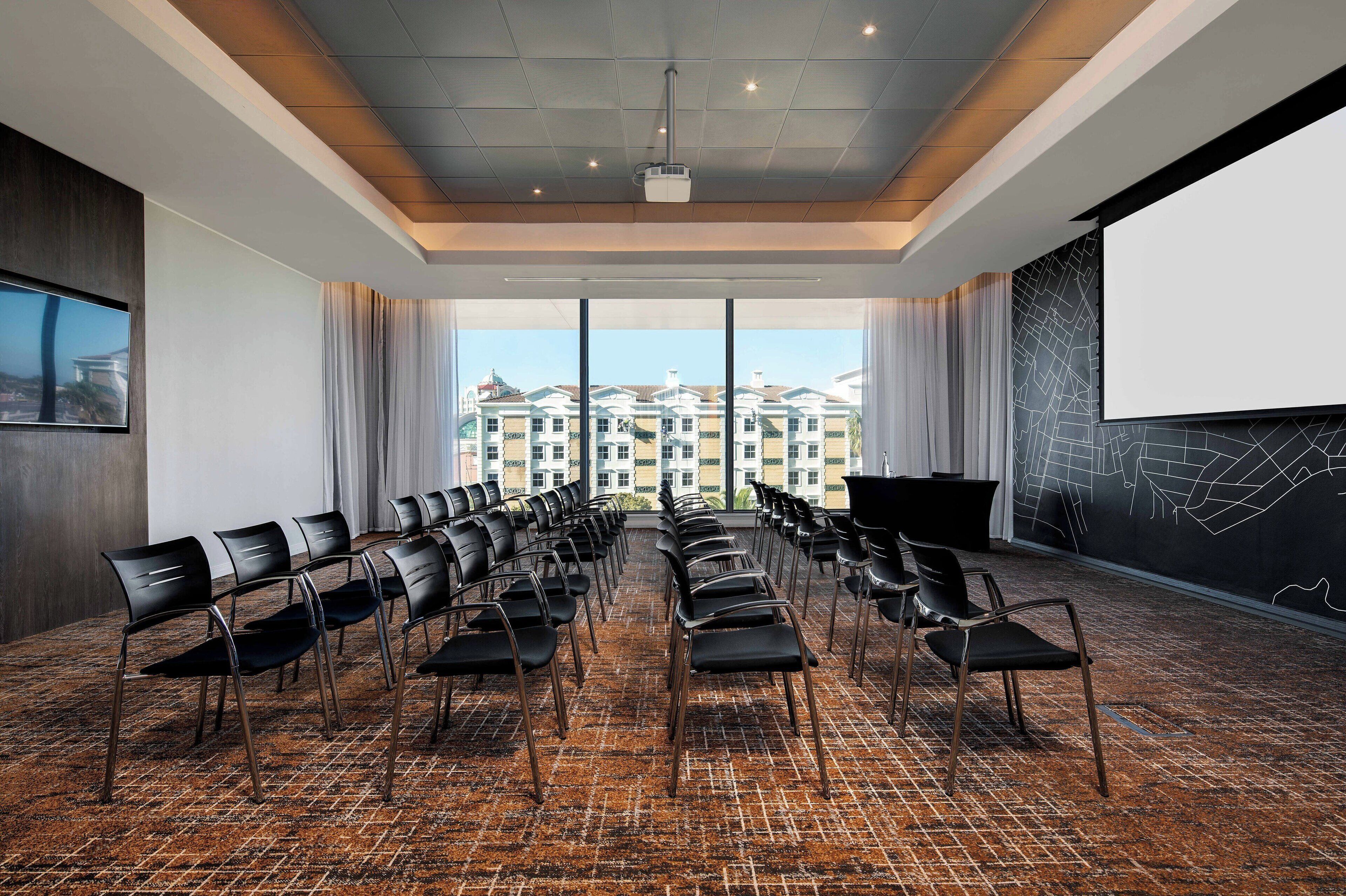 A conference room with rows of chairs, a projector screen, and a large window with a view of buildings.