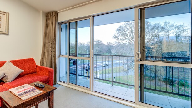 A room with a red sofa, a wooden coffee table with a magazine and remote control, and large windows offering a view of a street and trees. - Parkview On Hagley (Christchurch)
