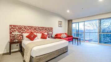 Un dormitorio con una cama grande, un sillón tapizado de color rojo, una mesita y una ventana que ofrece vistas a árboles y un vehículo rojo en el exterior.