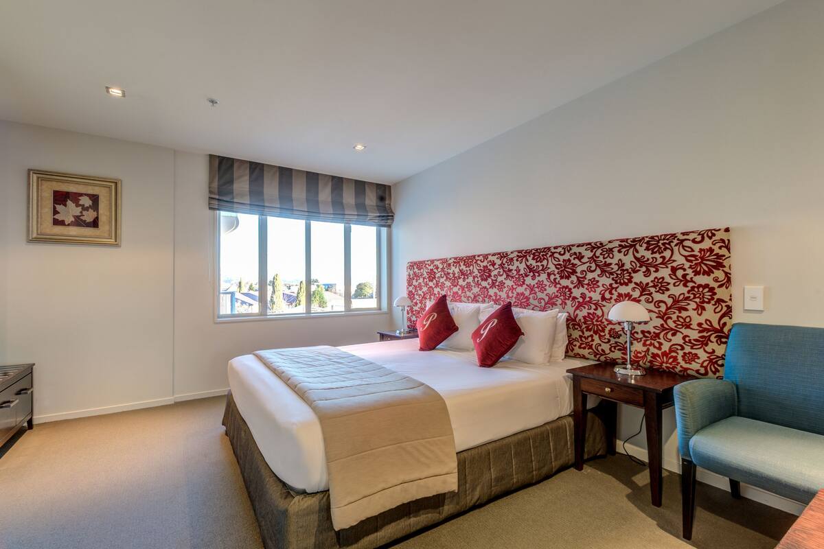 Une chambre à coucher avec un grand lit, une table de chevet, une chaise, une fenêtre donnant sur un beau paysage et une tête de lit à motifs floraux.