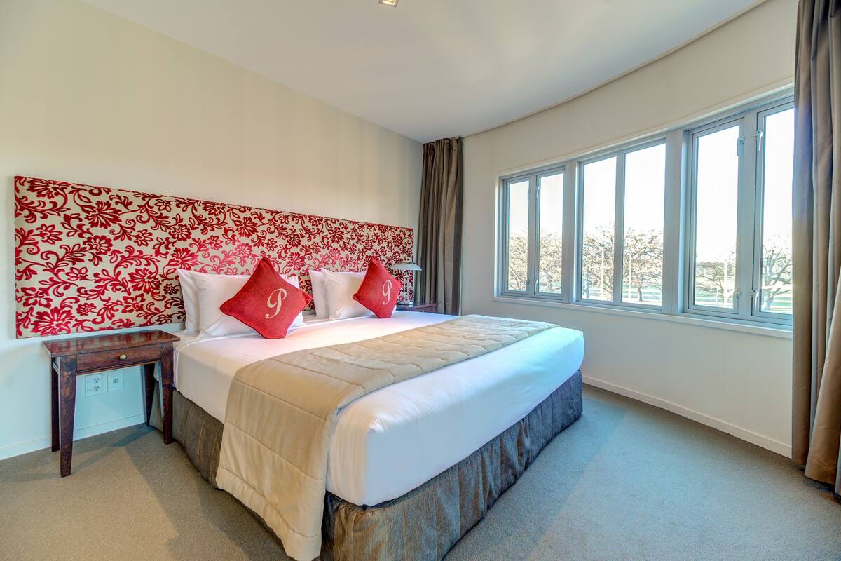 Une chambre à coucher avec un grand lit, une tête de lit ornée d’un motif floral et une fenêtre avec des rideaux.