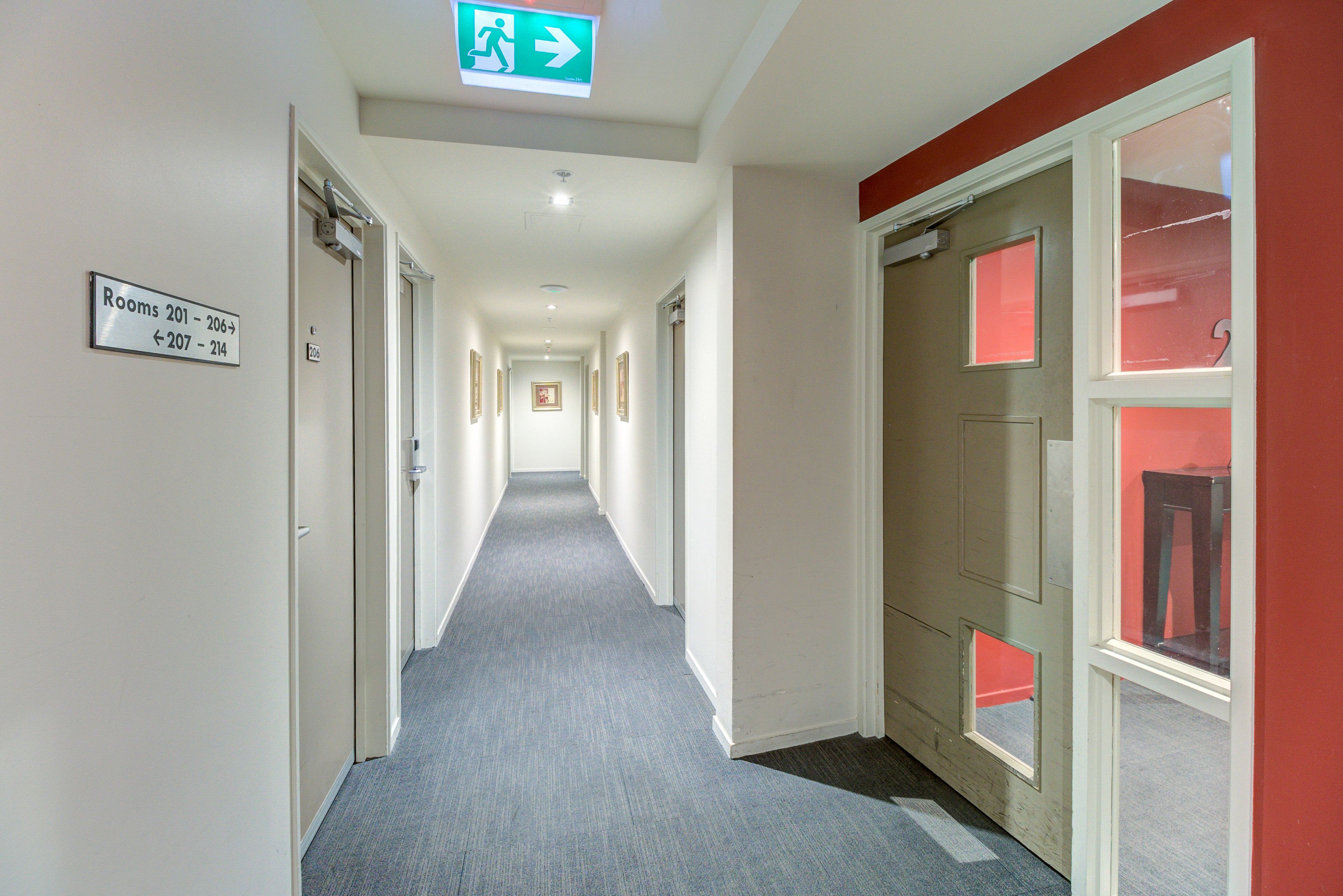 En hotelgang med skilte der viser vej til værelser 201-206 og 207-214, et skilt til nødudgang samt en dør med vindue i rødt ramme.