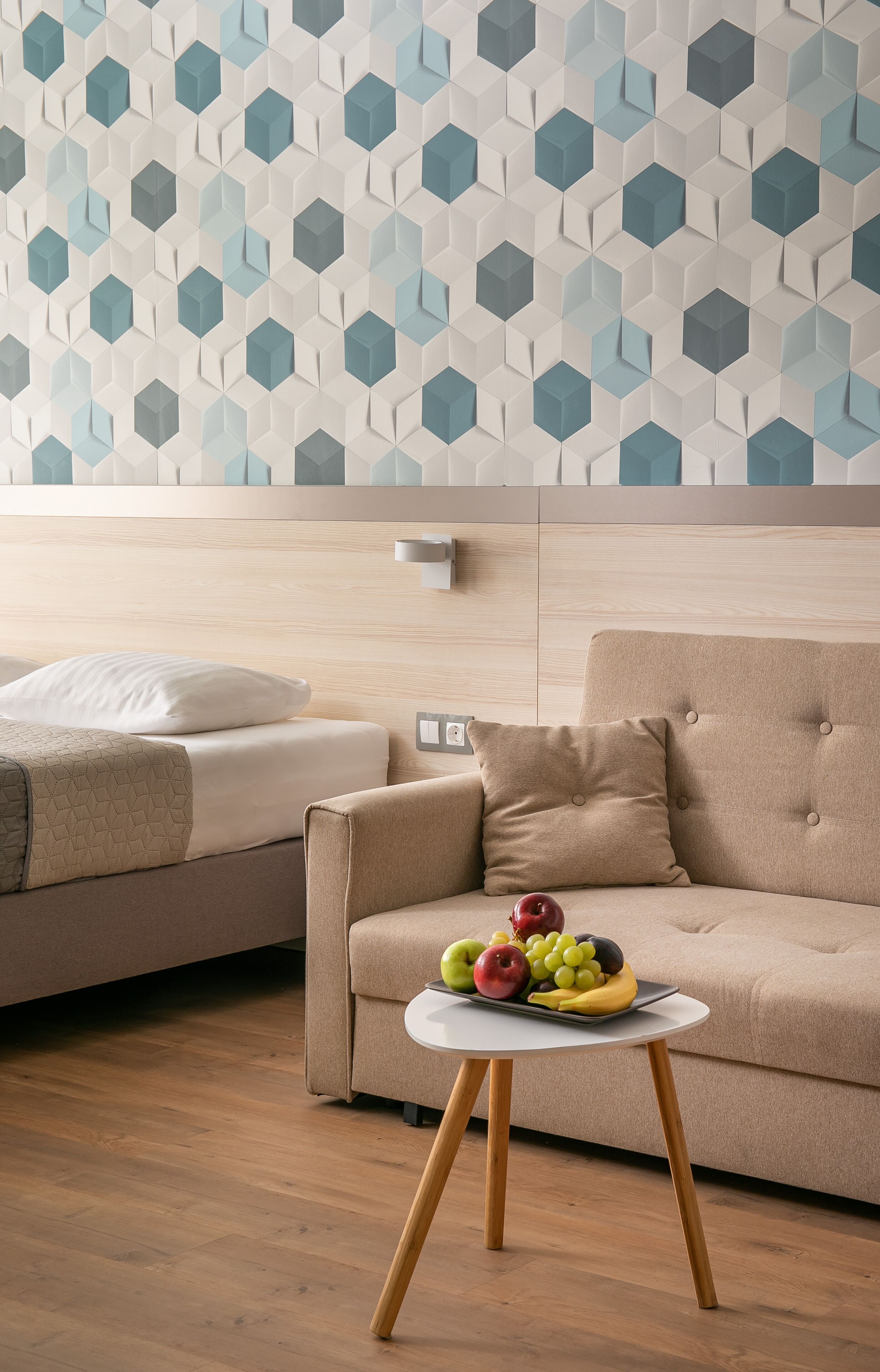Ein modernes Hotelzimmer mit Holzboden, einem beigefarbenen Sofa, einem weißen runden Tisch mit Obst und einer Wand mit geometrischem Muster.