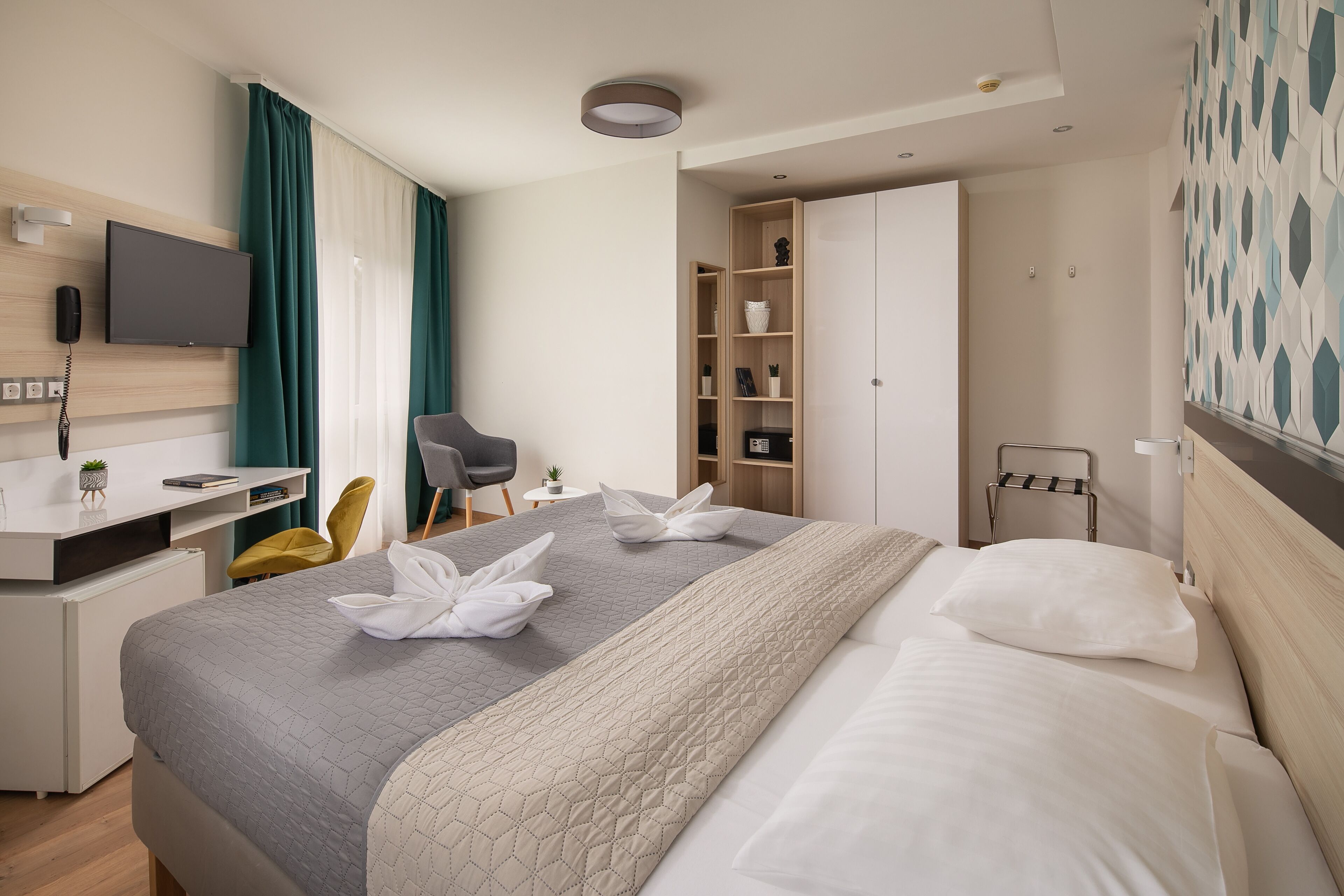 A modern hotel room with a neatly made bed, a desk with a computer, a chair, and a wardrobe.