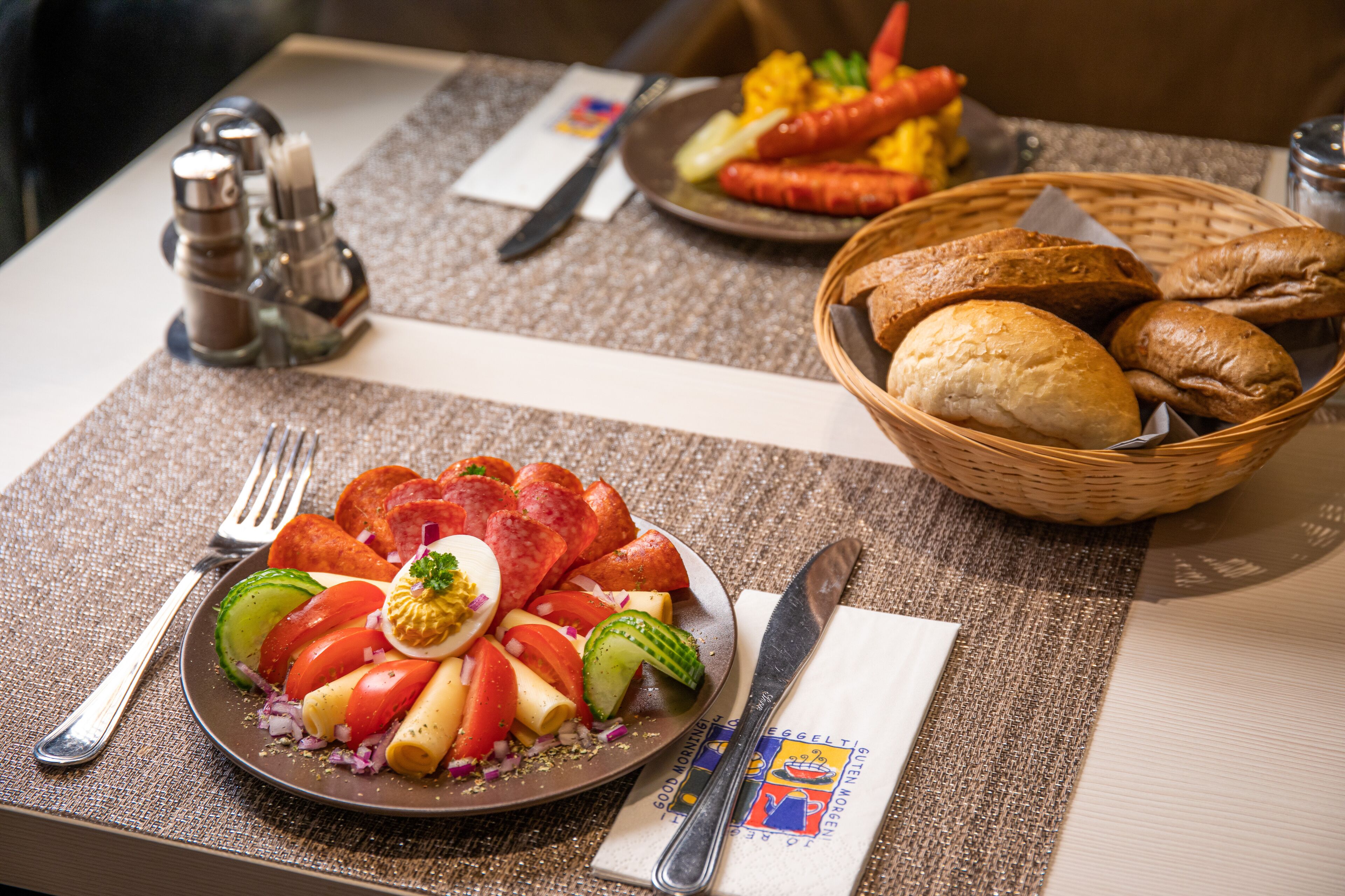 Un desayuno con un plato de verduras en rodajas, una cesta de pan y un plato de salchichas.