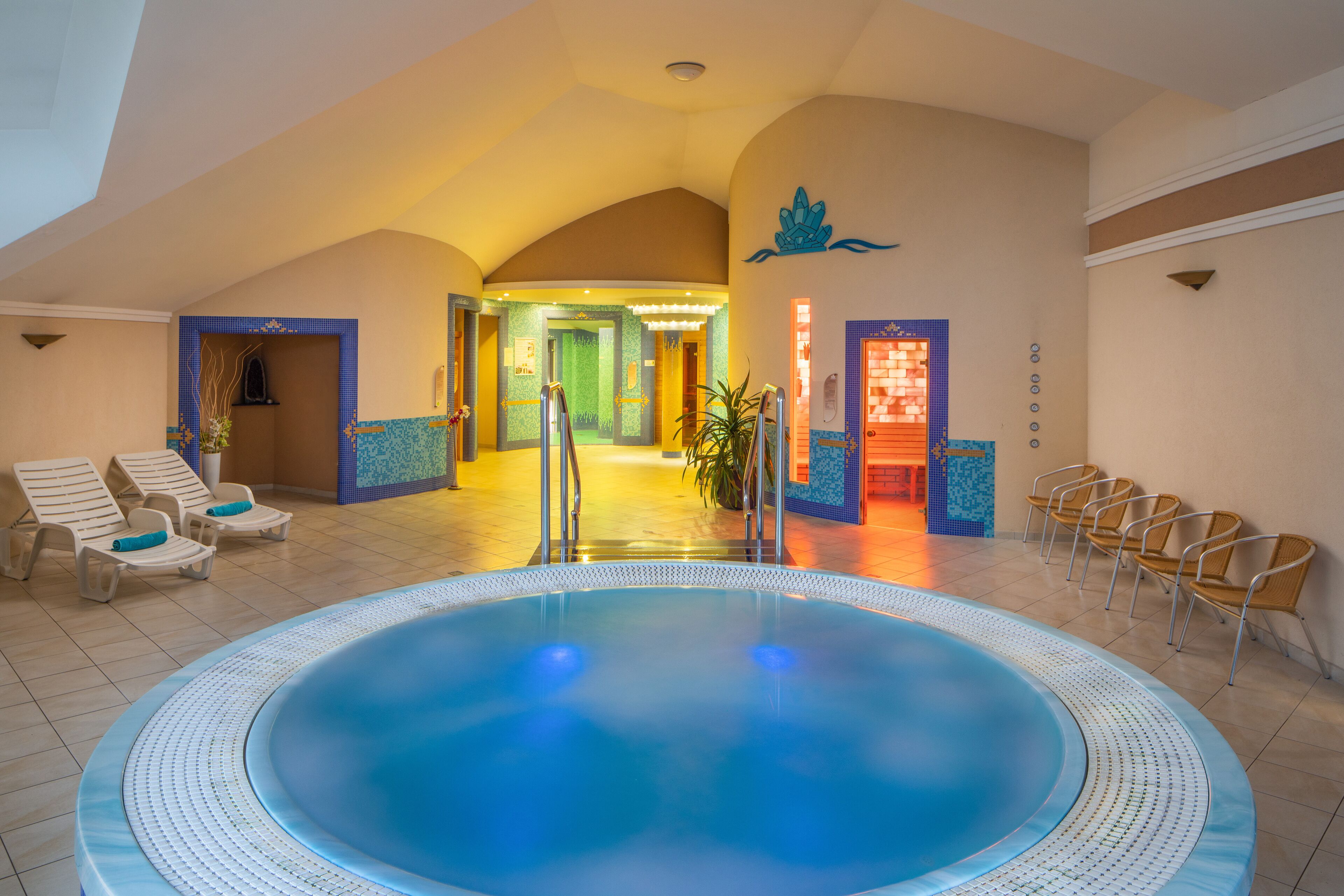 A large indoor swimming pool with a tiled deck, lounge chairs, and a reception area with a bright interior.