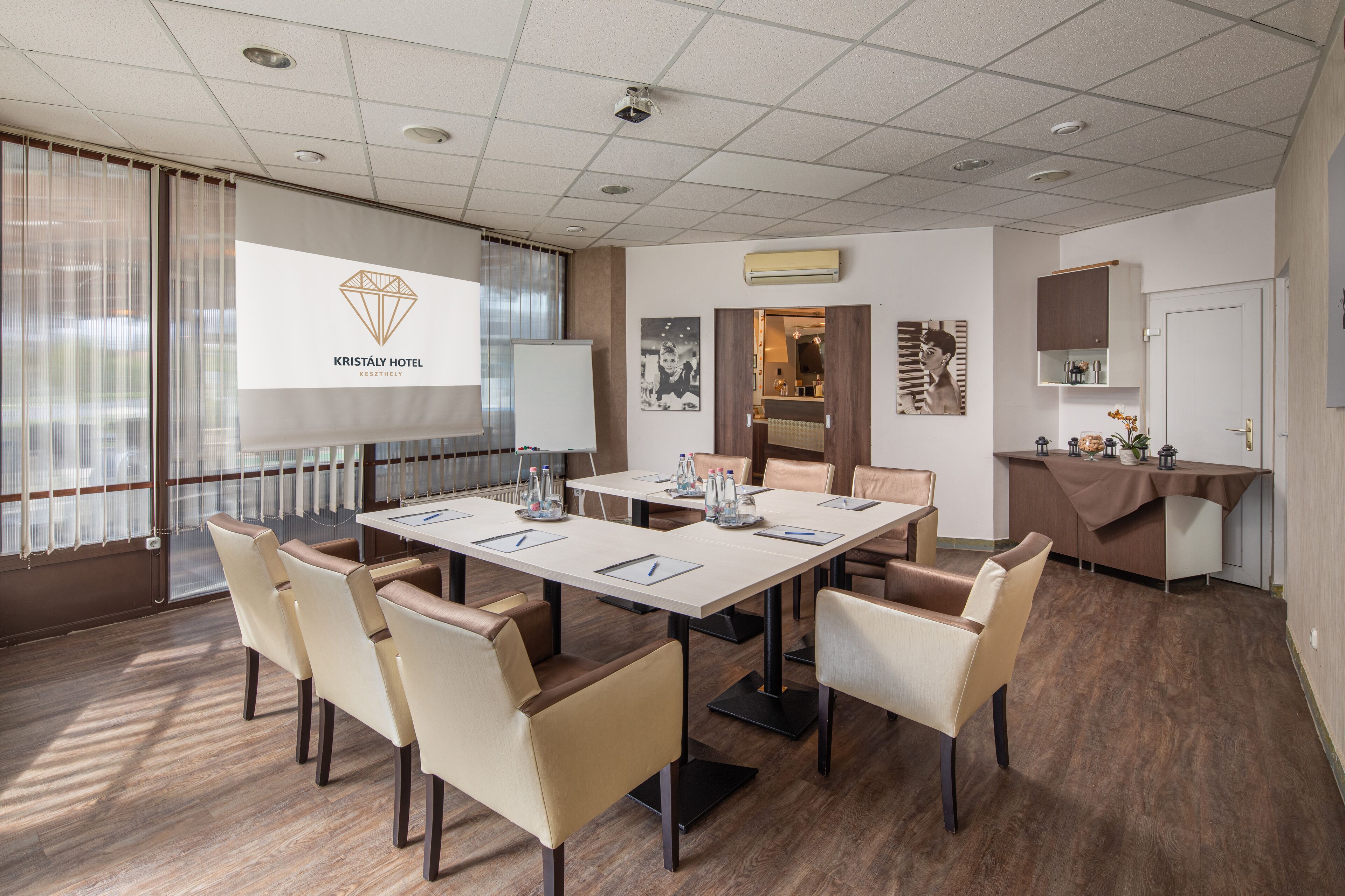 Un salón de conferencias con una amplia mesa, sillas, una pantalla de proyector mostrando un logotipo y una zona de cocina con mostrador y armarios.