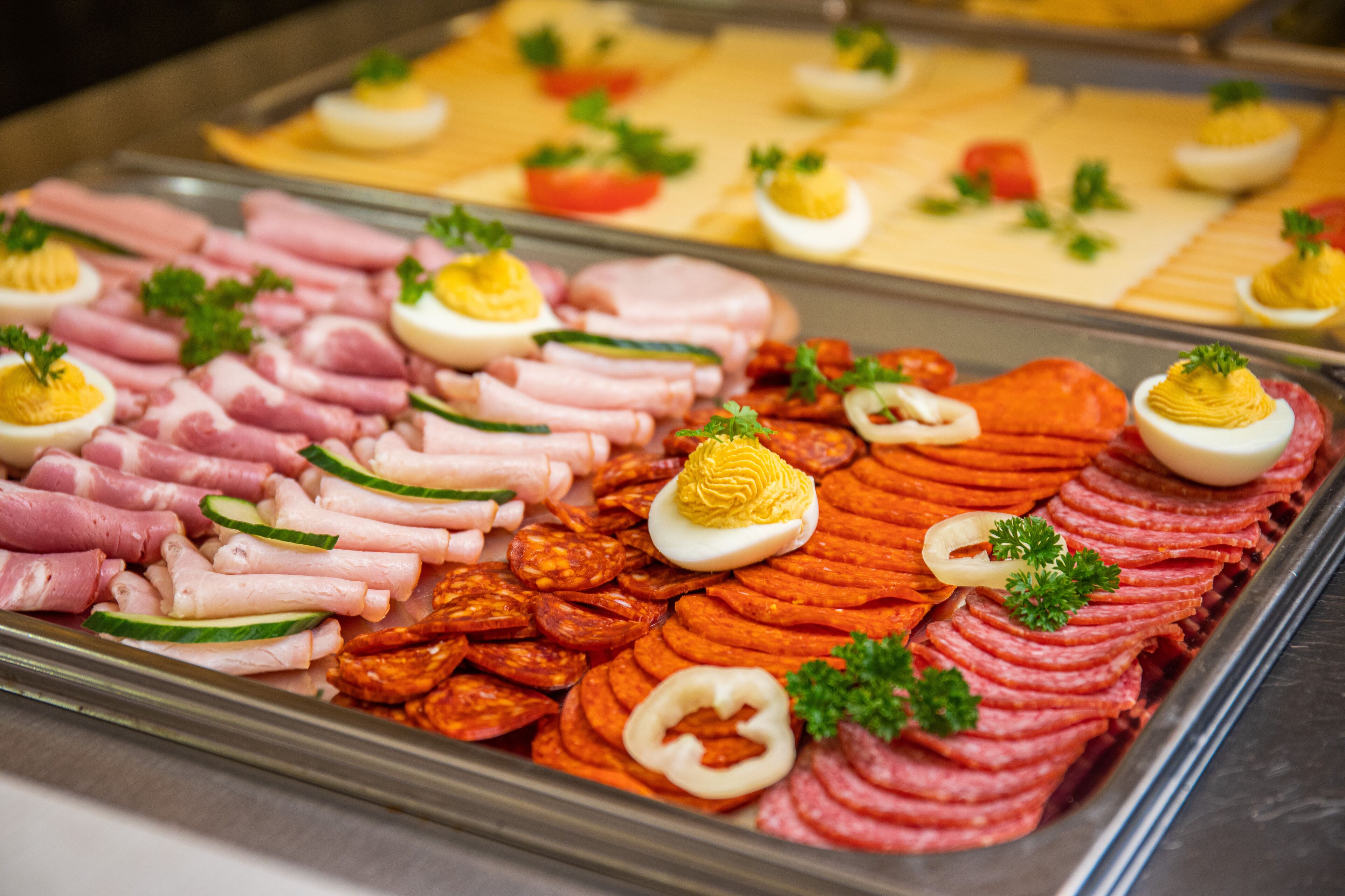 Un bufé con diversas carnes frías y embutidos, como jamón, tocino y salchichas, junto a guarniciones como perejil y lonchas de huevo.