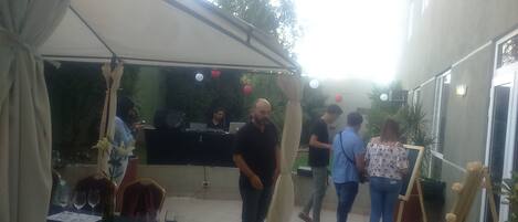 Uma área de pátio ao ar livre com mesas preparadas para refeições, um espaço para DJ e um grupo de pessoas reunidas informalmente.