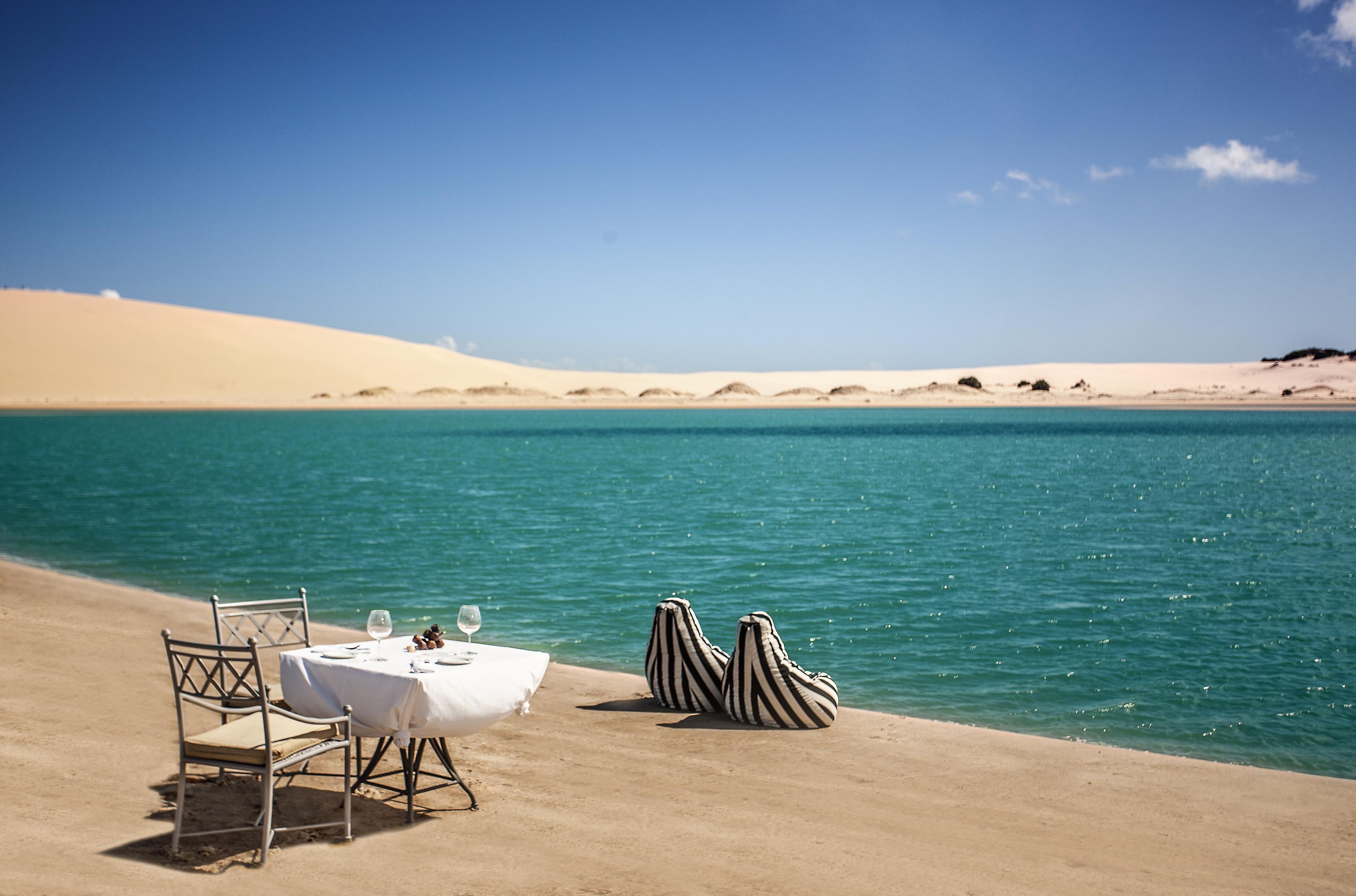 A dining setup with a table and chairs on a sandy beach, overlooking a calm sea and distant dunes.