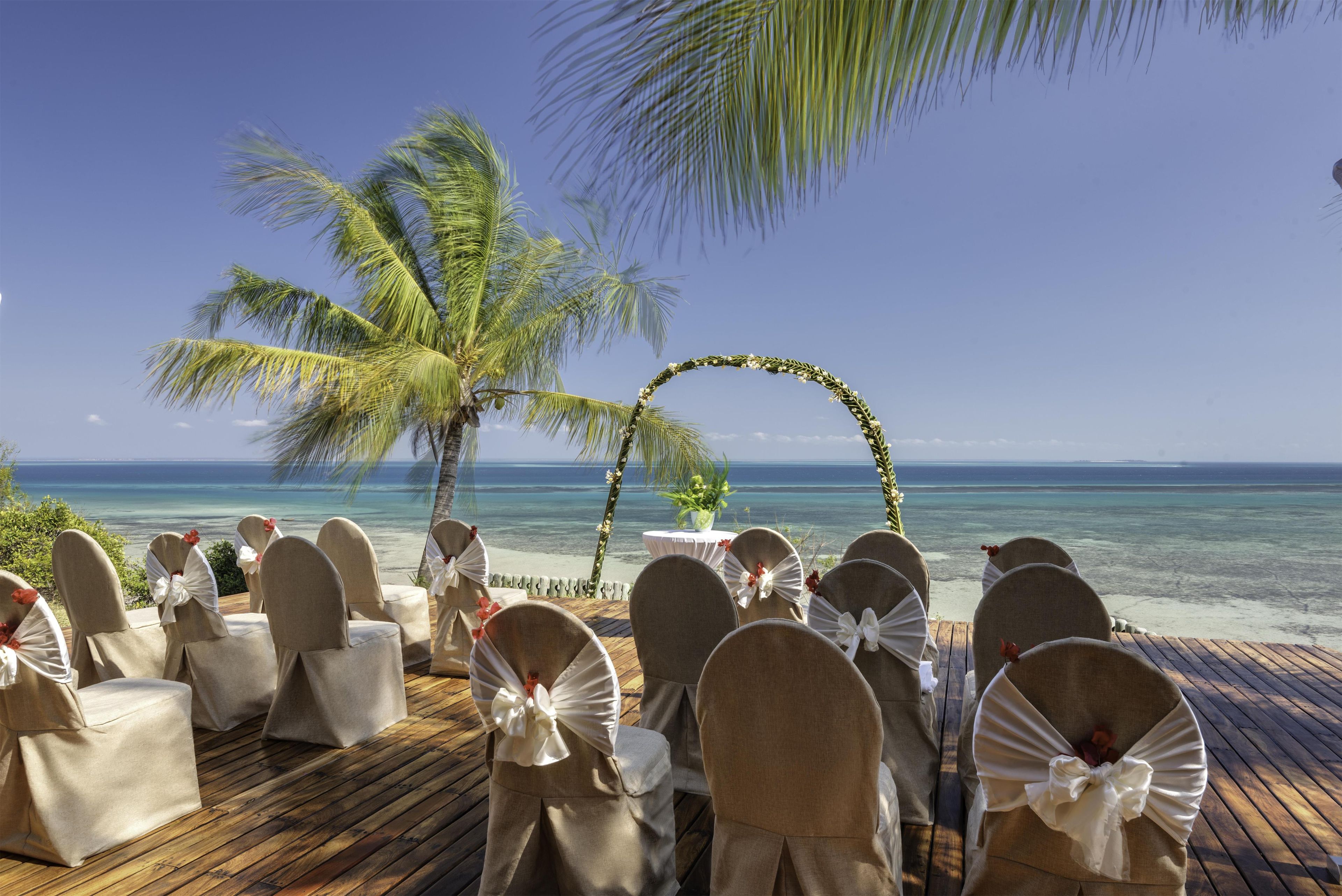 A beachside wedding setup with a wooden deck, chairs with white bows, a floral arch, and a view of the ocean.