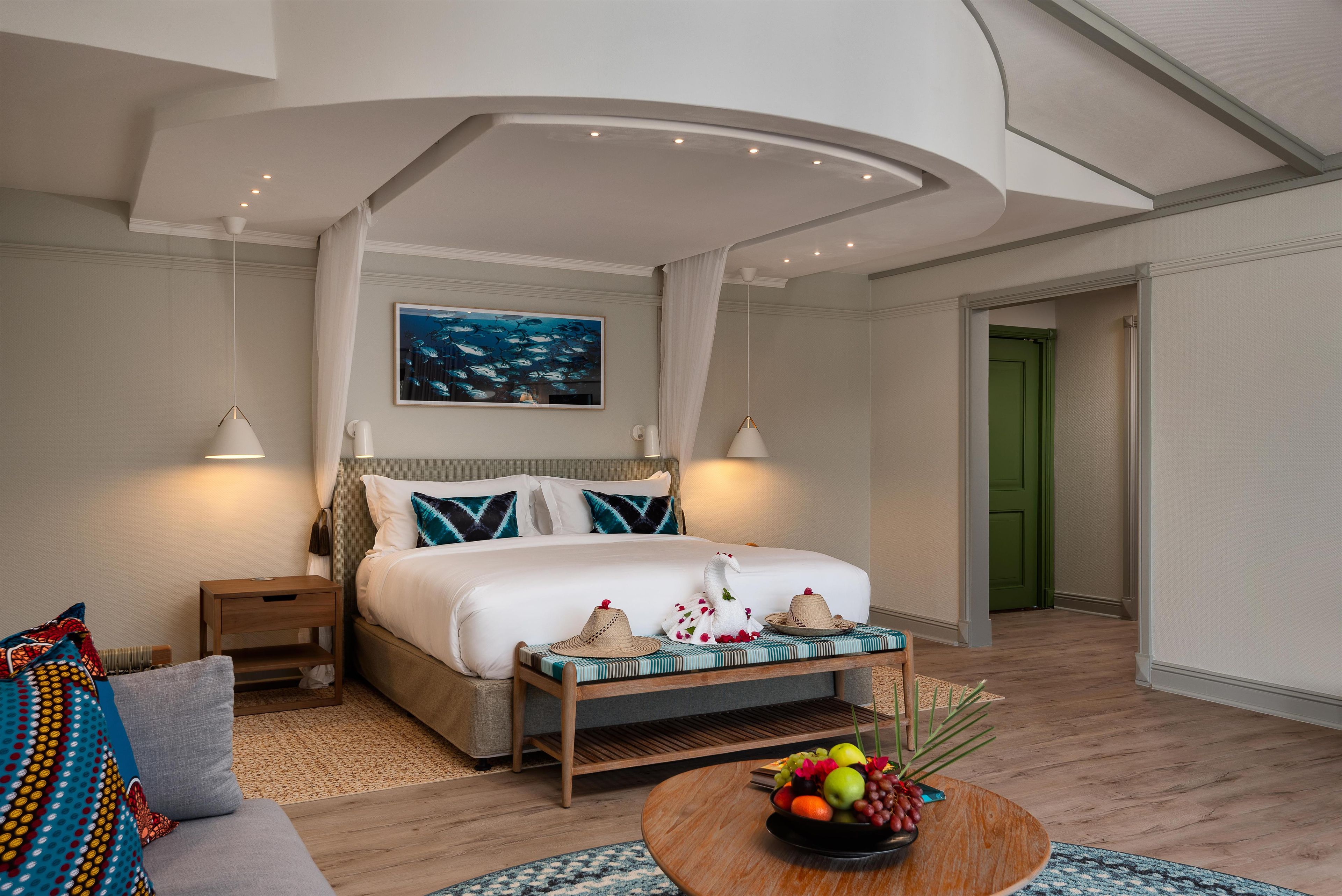 Une chambre d’hôtel avec un lit, des tables de chevet, une petite table de nuit, un tableau accroché au mur, une porte verte et un panier de fruits posé sur une table en bois.