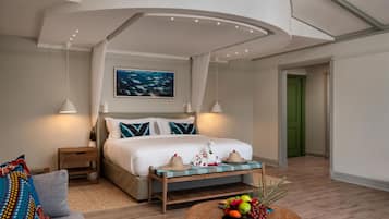Une chambre d’hôtel avec un lit, des tables de chevet, une petite table de nuit, un tableau accroché au mur, une porte verte et un panier de fruits posé sur une table en bois.
