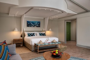 Habitación de hotel con cama, mesitas de noche, un cuadro colgado en la pared, una puerta verde y un frutero sobre una mesa de madera.