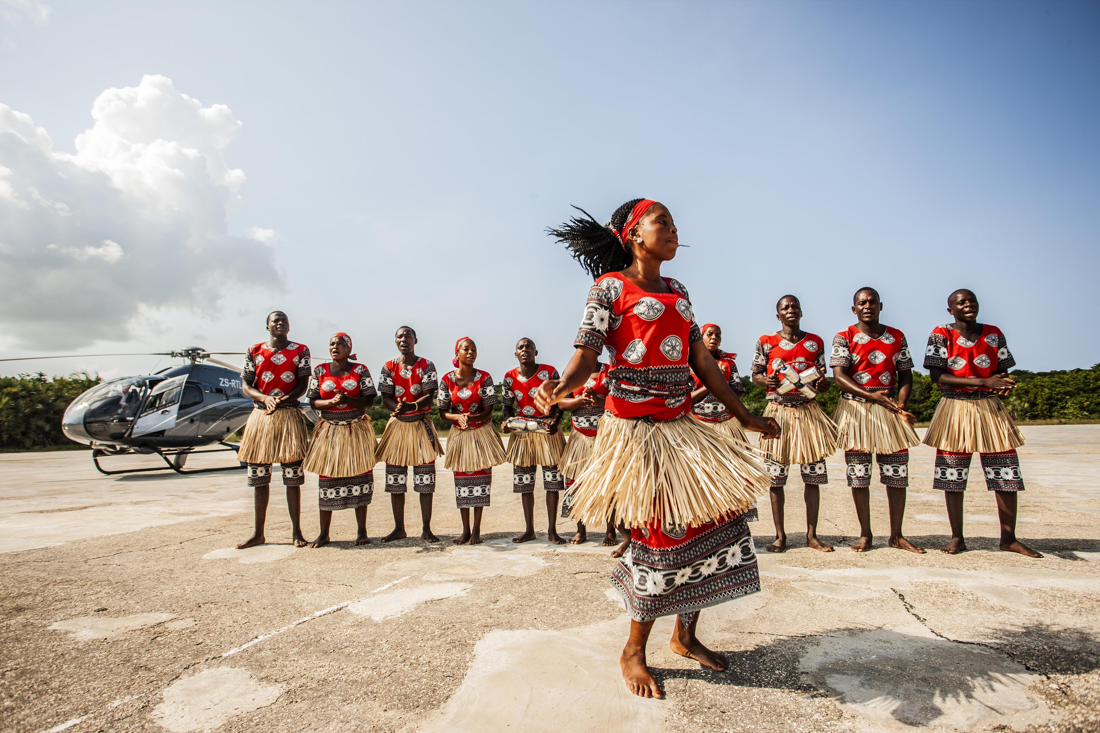 Un grupo de personas vestidas con atuendos tradicionales sobre una superficie de concreto, con un helicóptero al fondo.