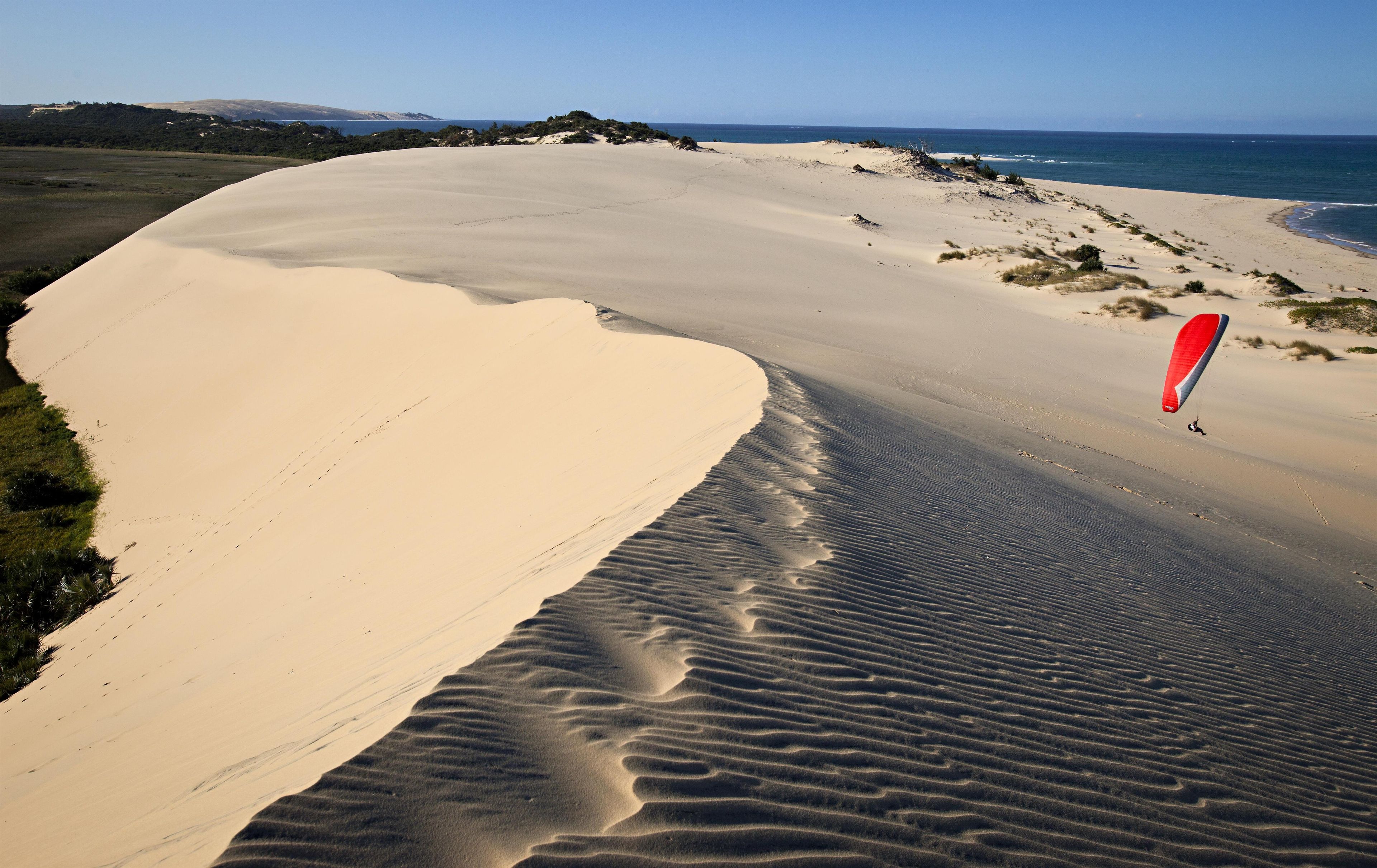 A beach with sand dunes and a person paragliding.