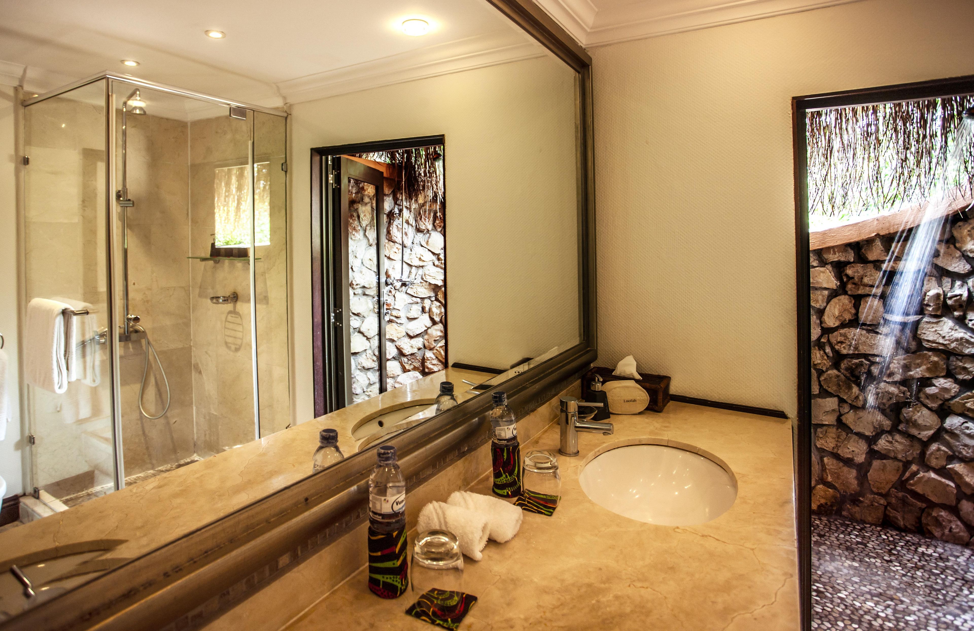 Bagno con doccia vetrata, uno specchio grande, un lavandino con articoli da toilette e una parete in pietra a vista.