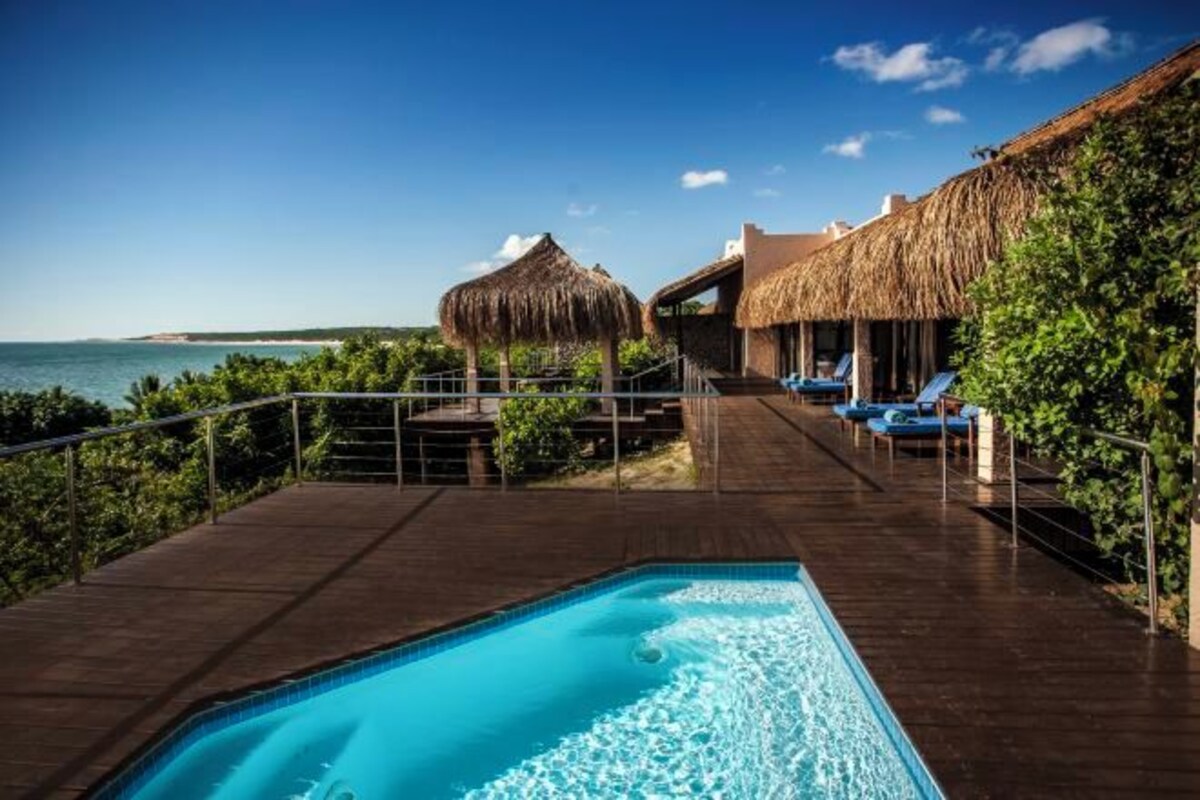 Un espace piscine avec vue sur l’océan, une structure à toit de chaume et des chaises longues.