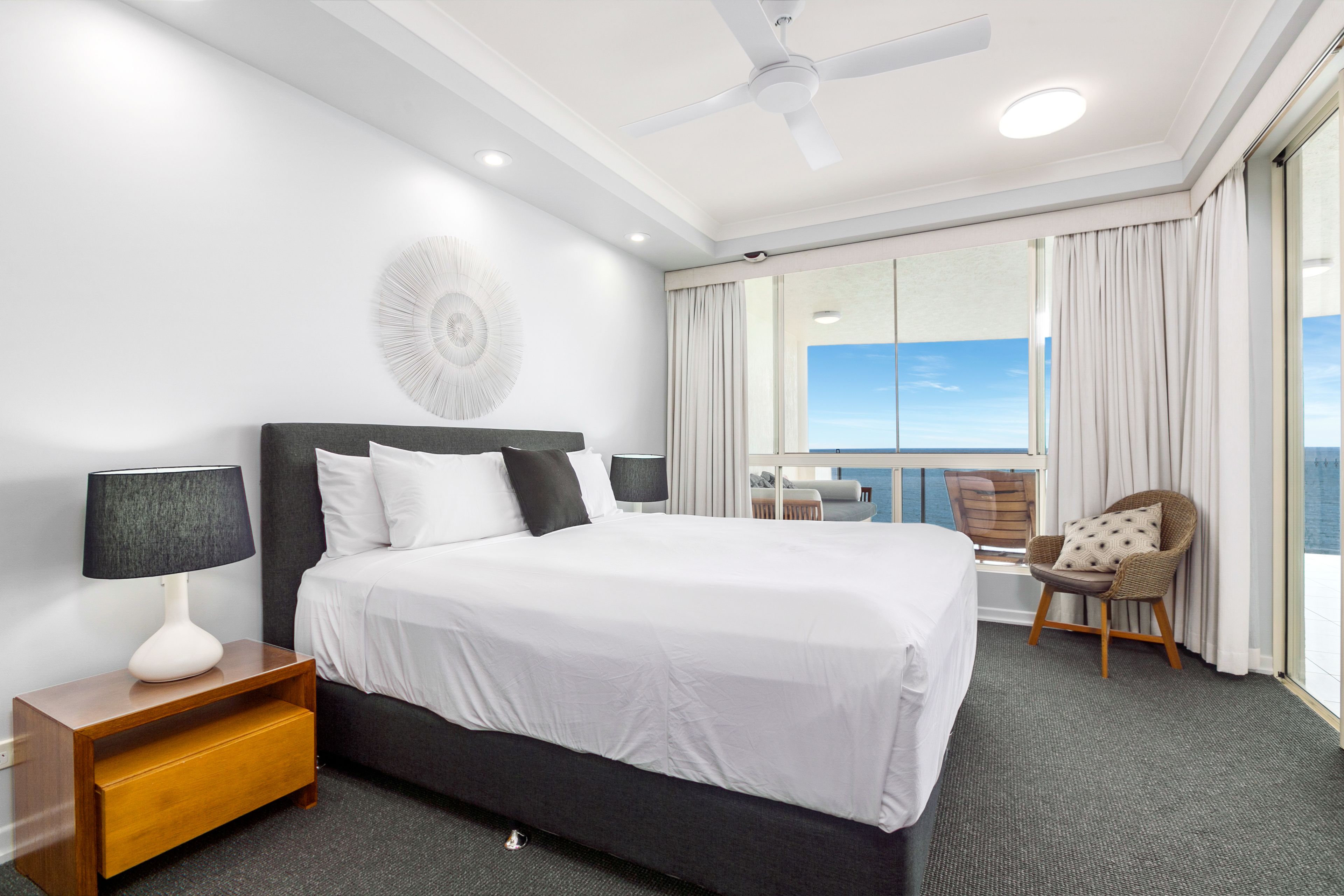 Une chambre d’hôtel avec un grand lit, une table de chevet avec une lampe, une chaise et une vue sur le ciel par la fenêtre.