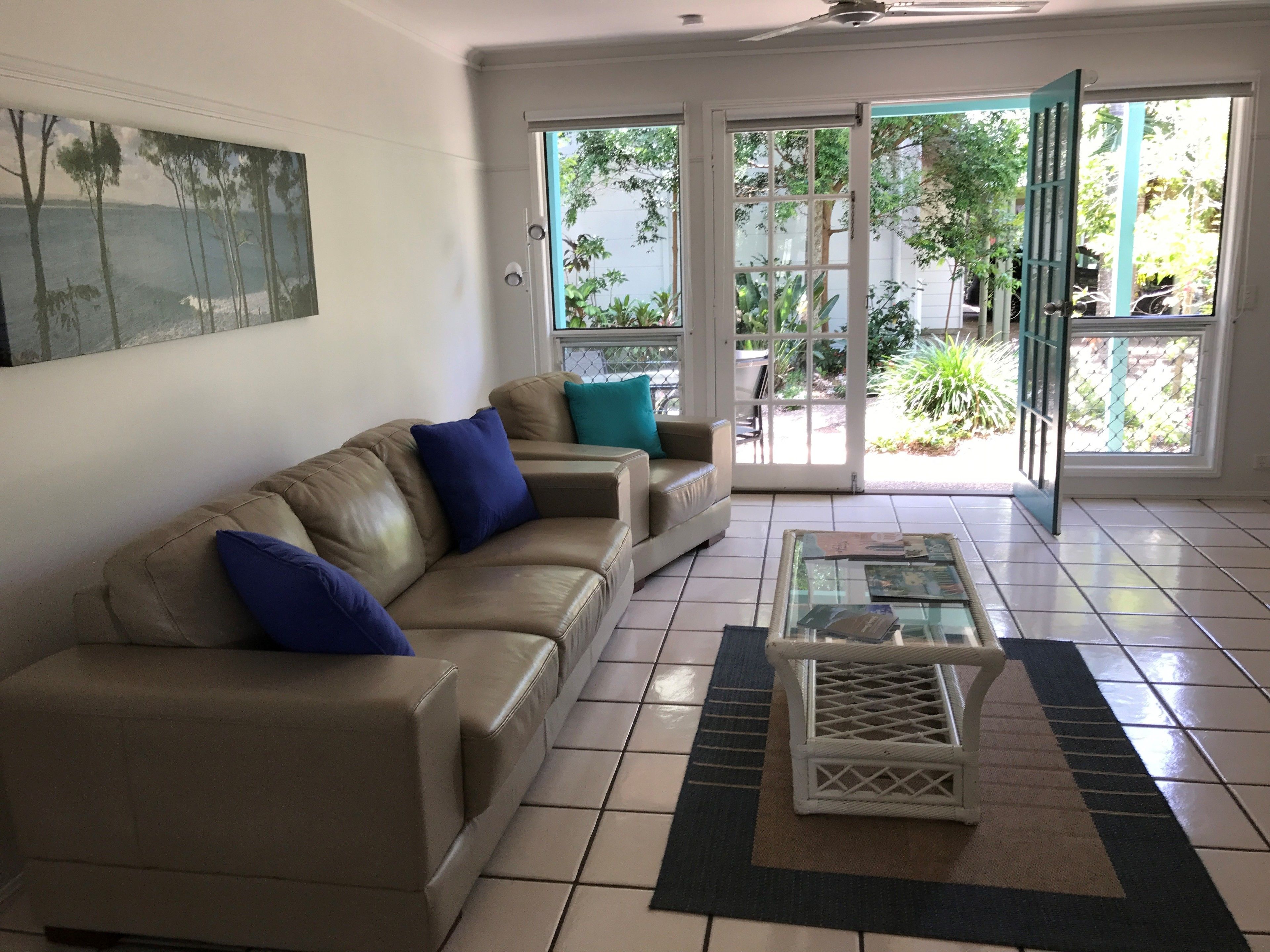 A living room with a beige sofa, blue cushions, a glass coffee table, and a view of a garden through glass doors.