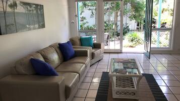 A living room with a beige sofa, blue cushions, a glass coffee table, and a view of a garden through glass doors.