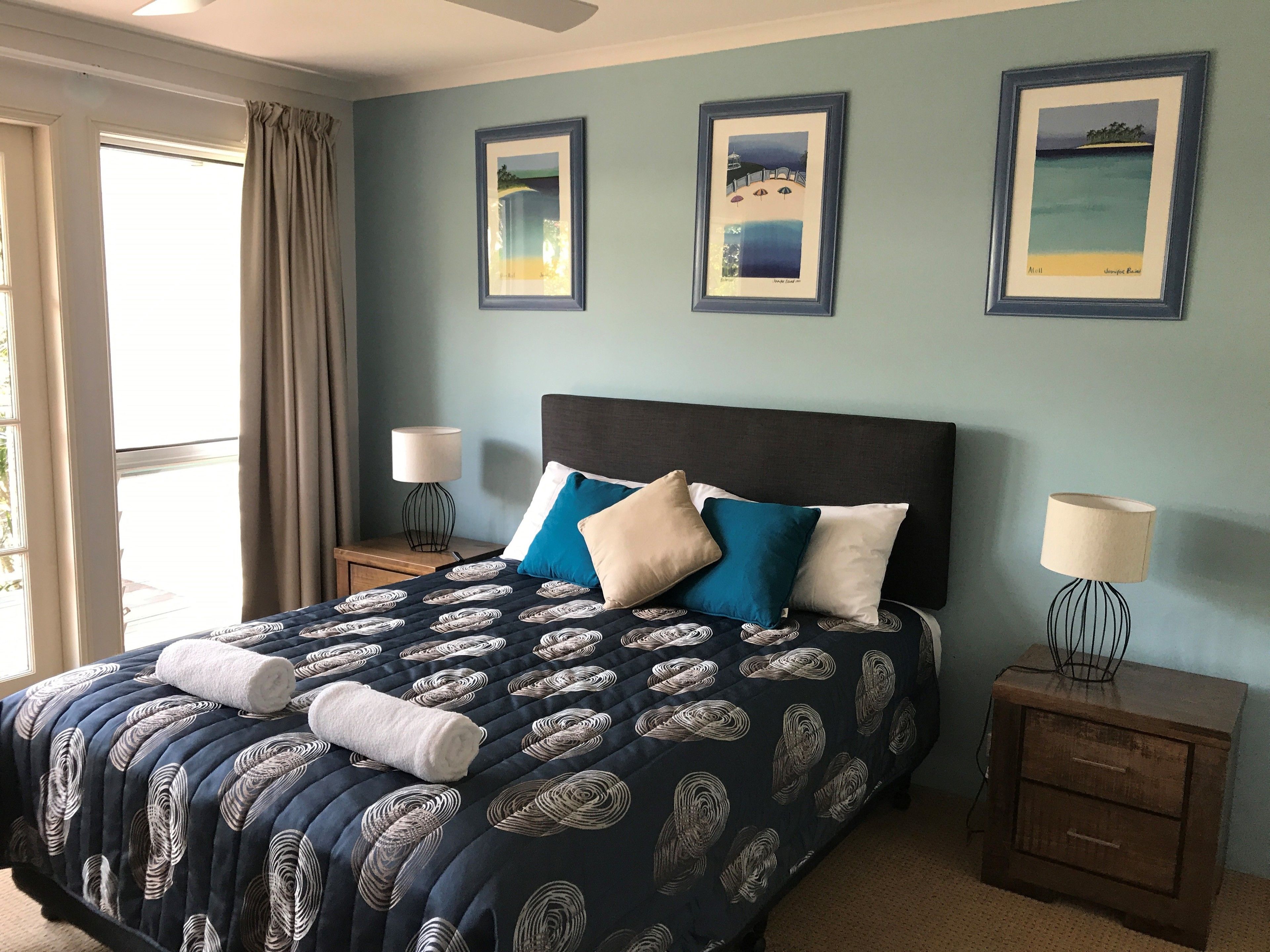 A bedroom with a bed, two bedside tables, a window with curtains, and three framed pictures on the wall.
